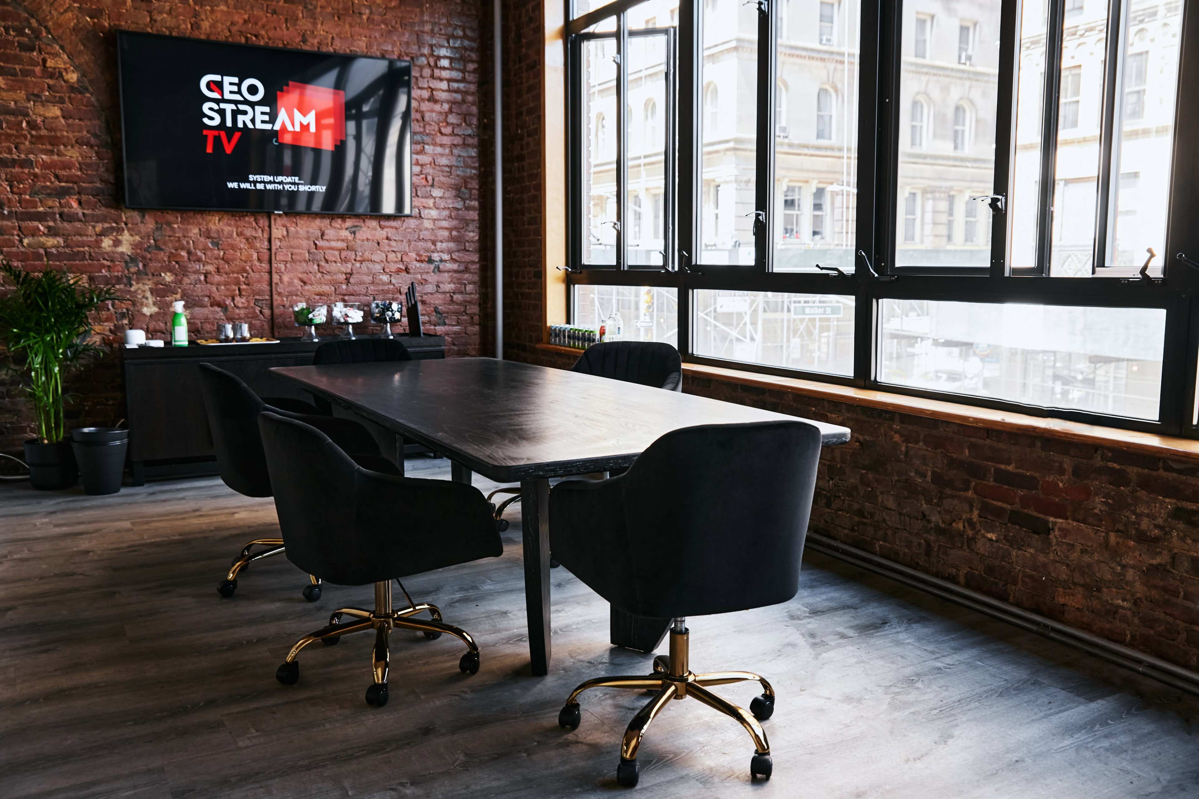 A modern conference room features a large table surrounded by black swivel chairs, with a television displaying "CEO STREAM TV" on a brick wall.