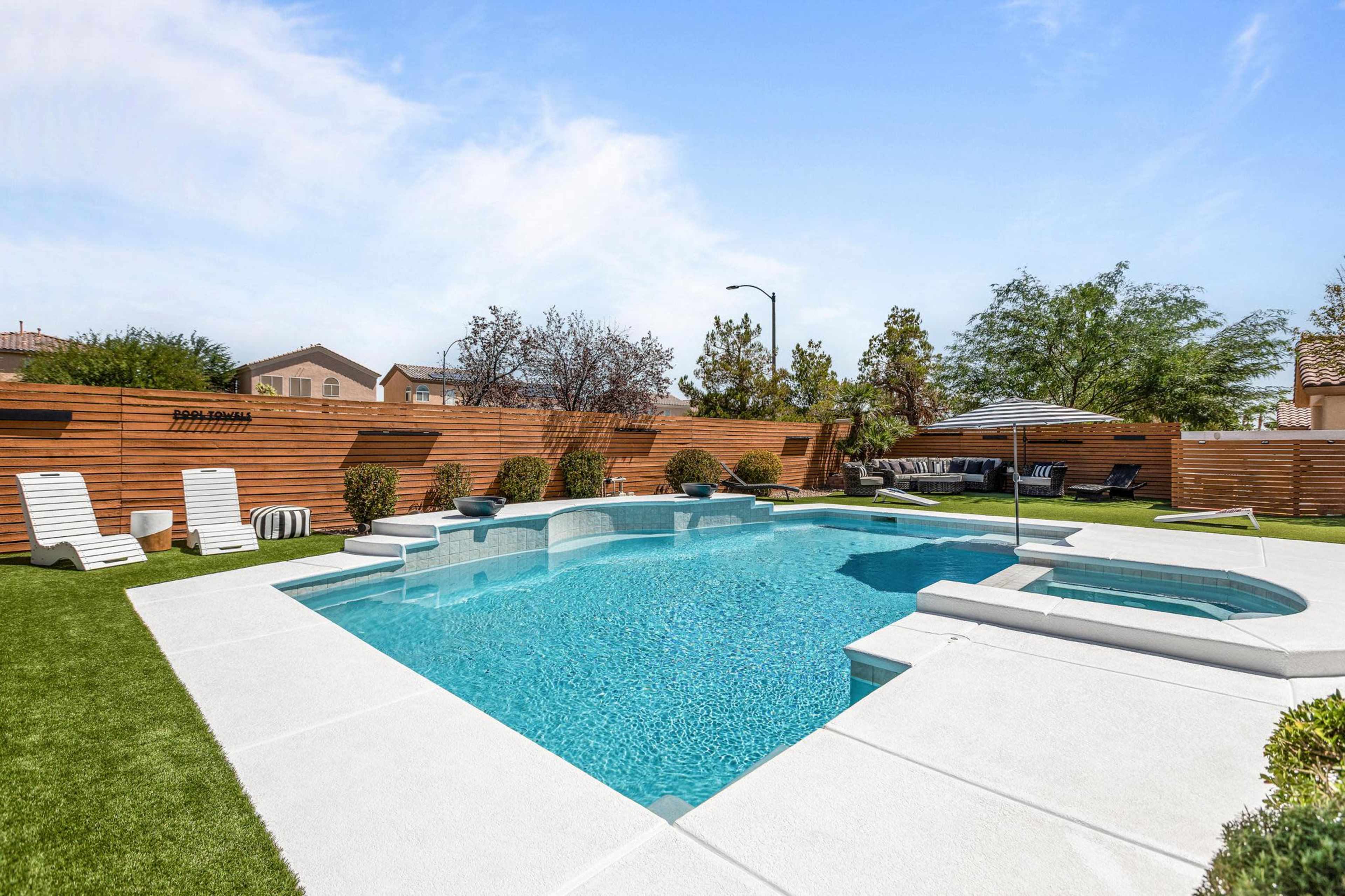 The image shows a backyard with a swimming pool, spa, and lounge area surrounded by wooden fencing and landscaped greenery.