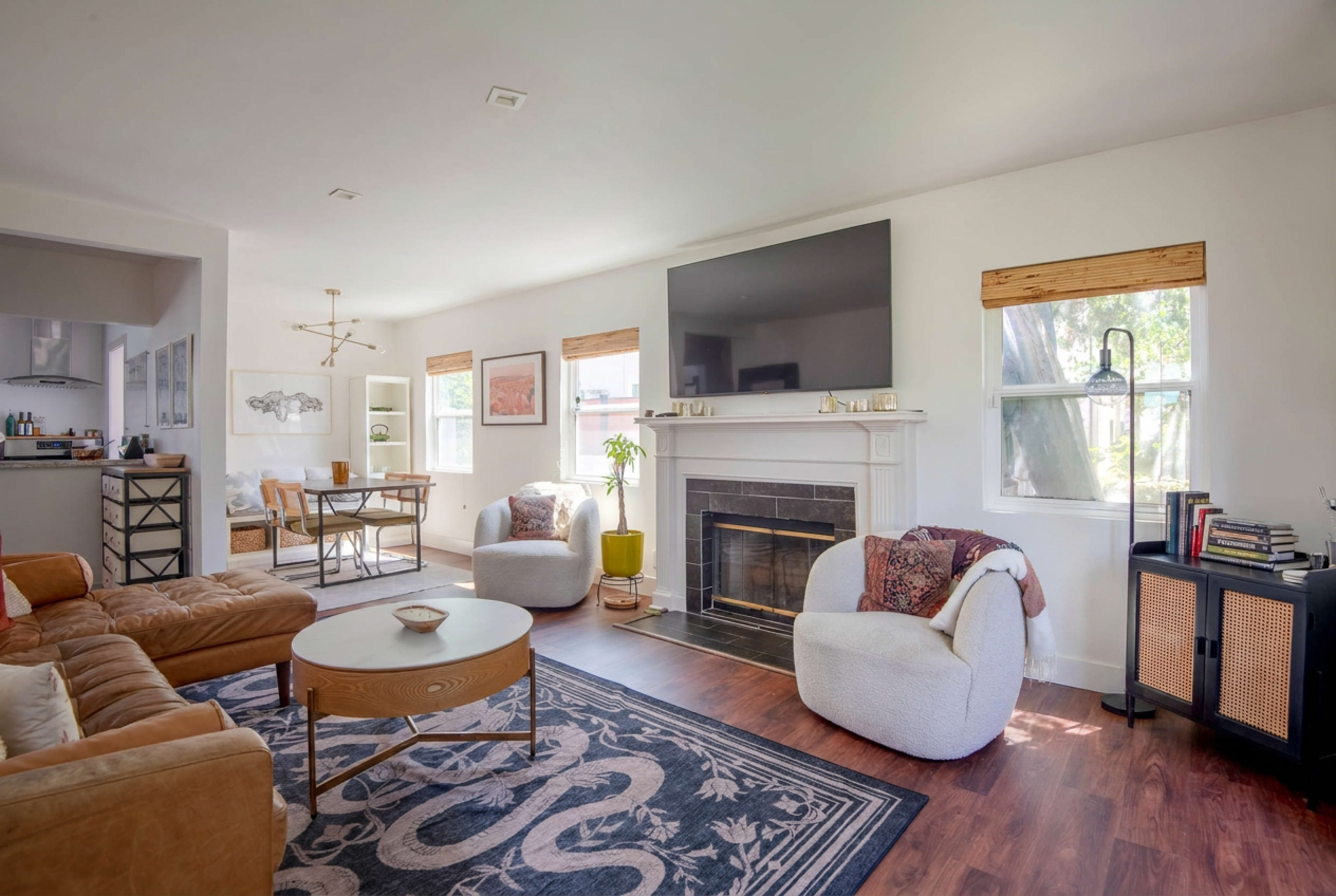 The image depicts a bright living room with a central fireplace, two armchairs, a sofa, a coffee table, and a large wall-mounted television.