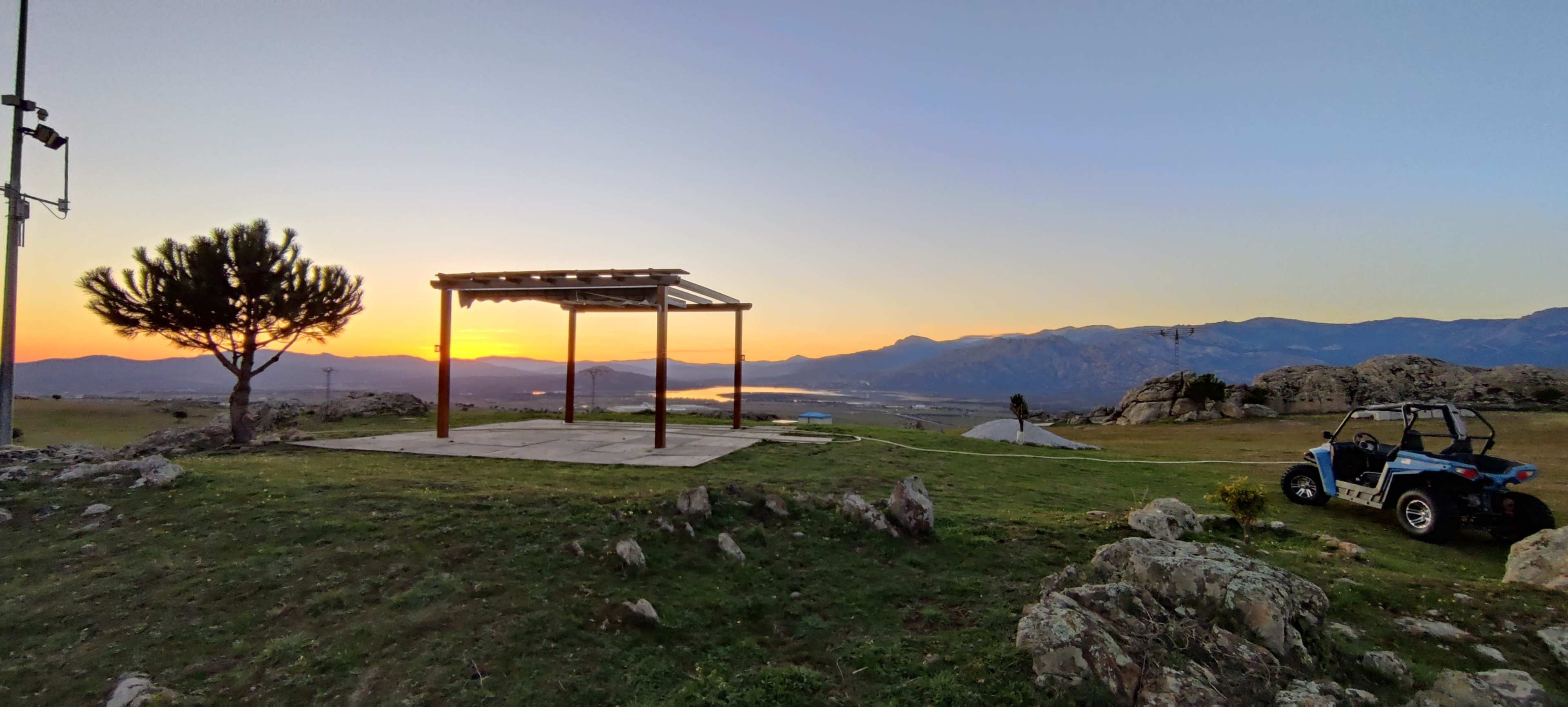 A wooden gazebo stands on a grassy area overlooking a sunset with mountains in the background.