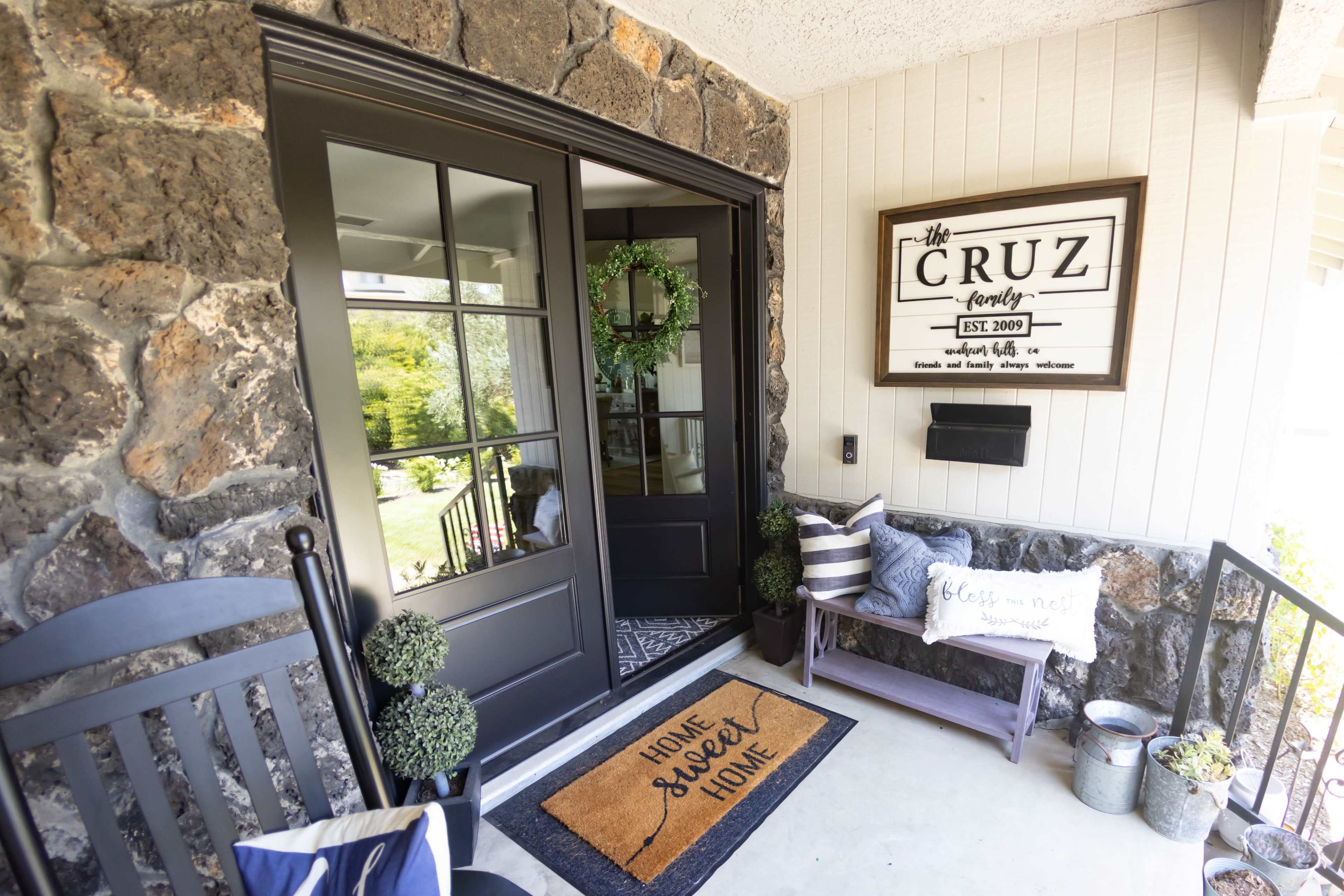 A front porch with stone walls, a dark double door, a decorative welcome mat, and various potted plants.