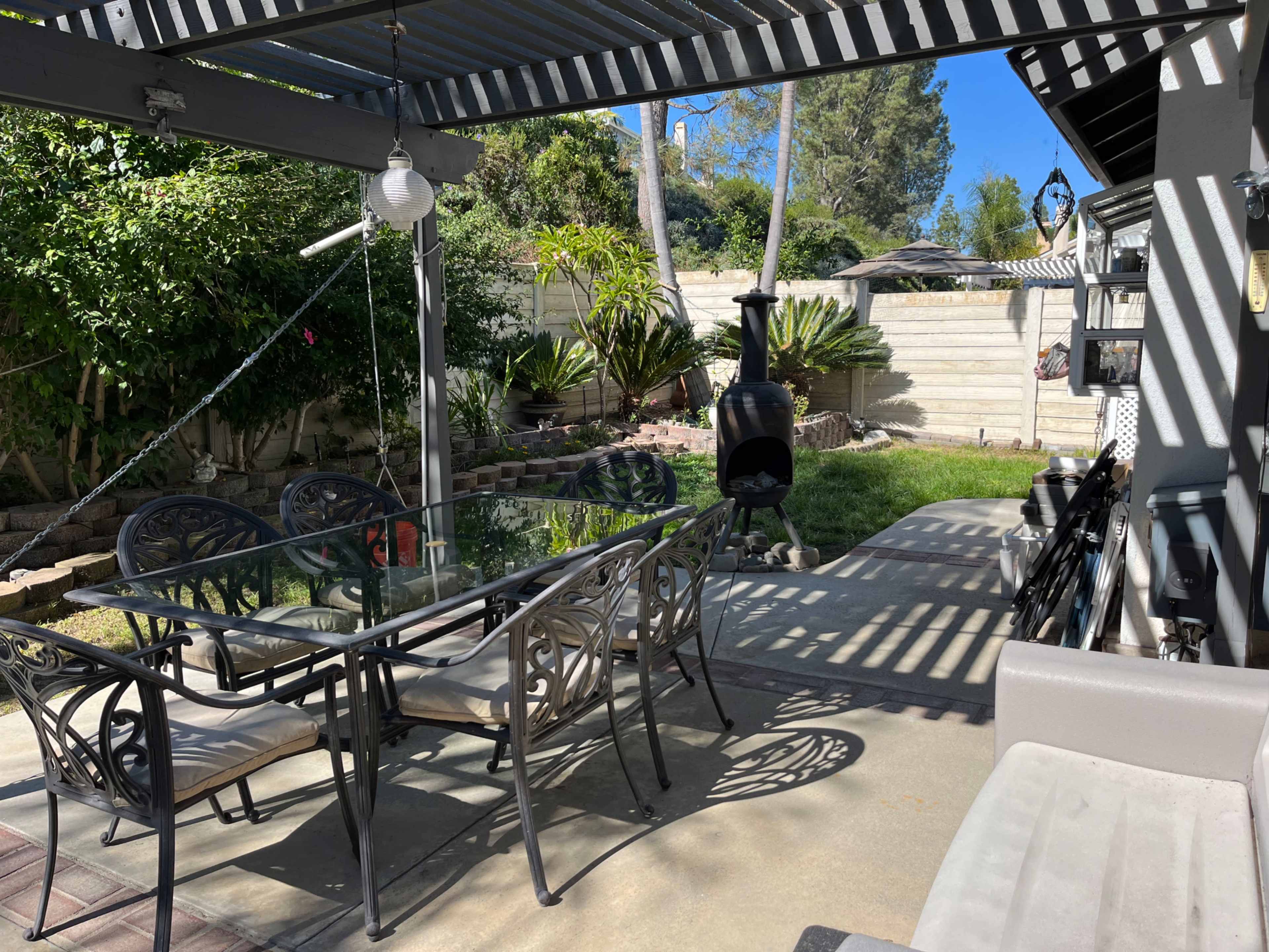 A patio area features a glass table with black wrought iron chairs, surrounded by greenery and a black outdoor heater.
