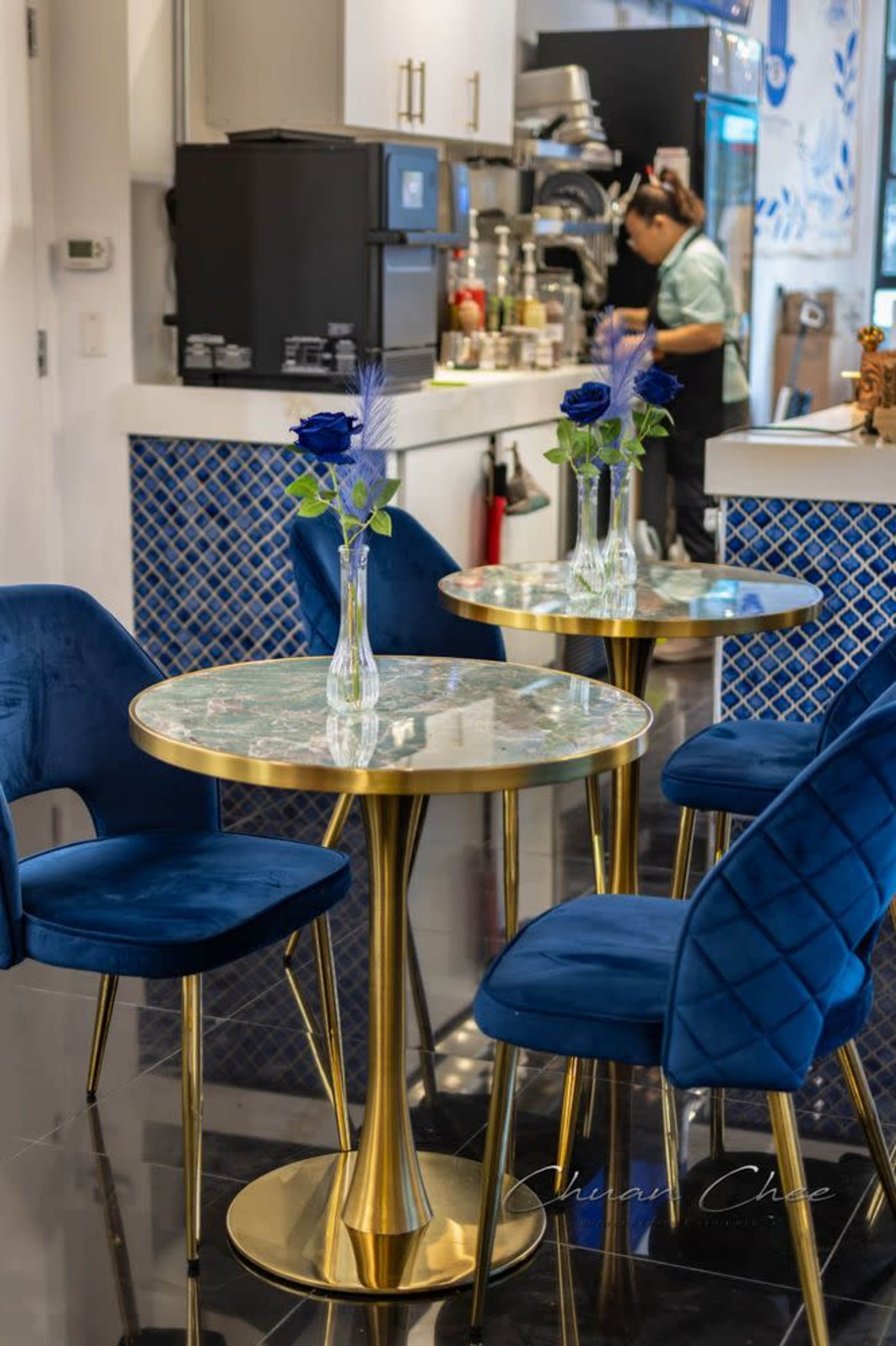 The image shows a modern café interior with blue chairs and a marble-top table featuring vases with blue roses.