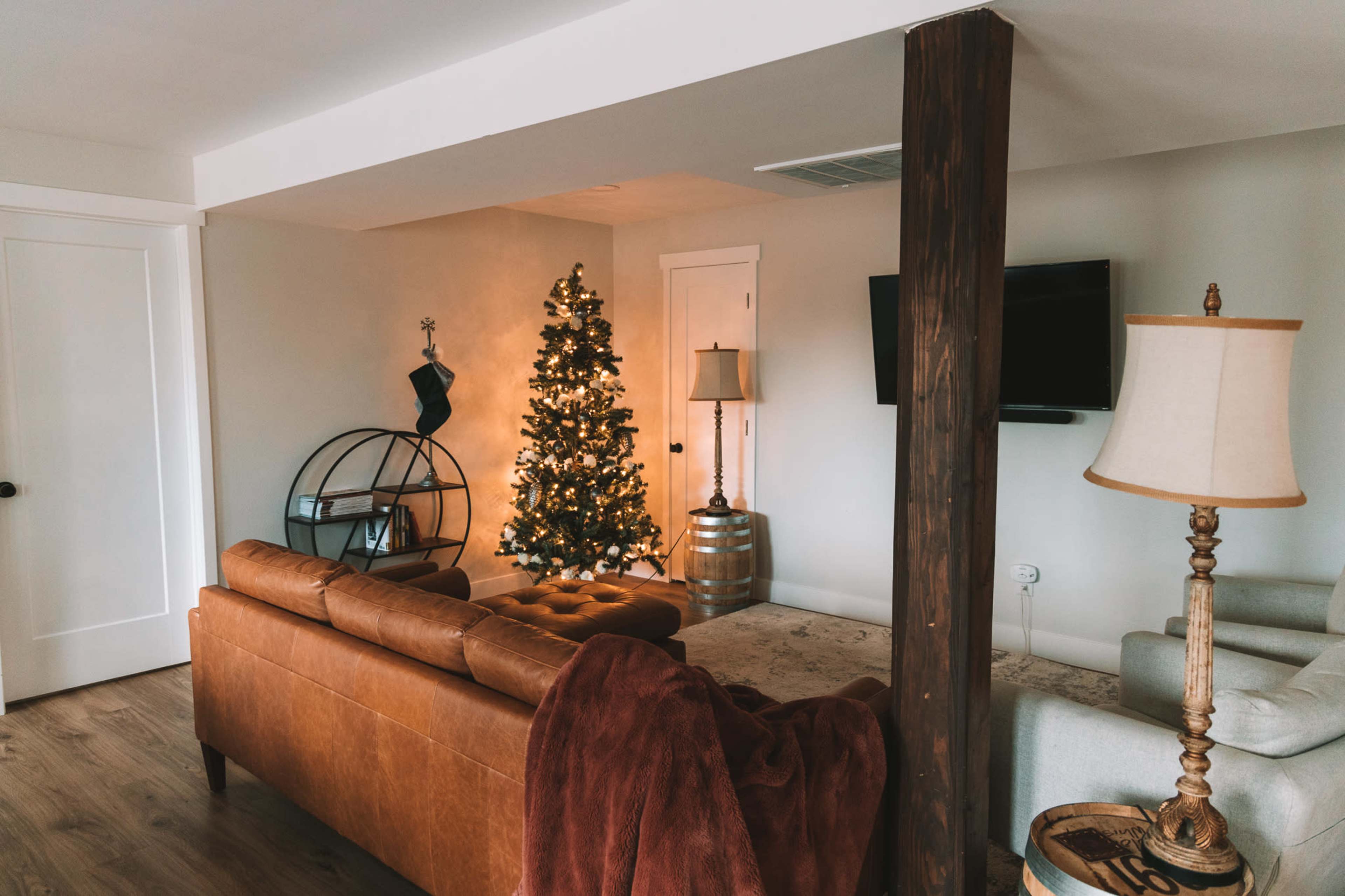 A cozy living room featuring a leather sofa, a decorated Christmas tree, and two lamps, with a wooden column separating the space.