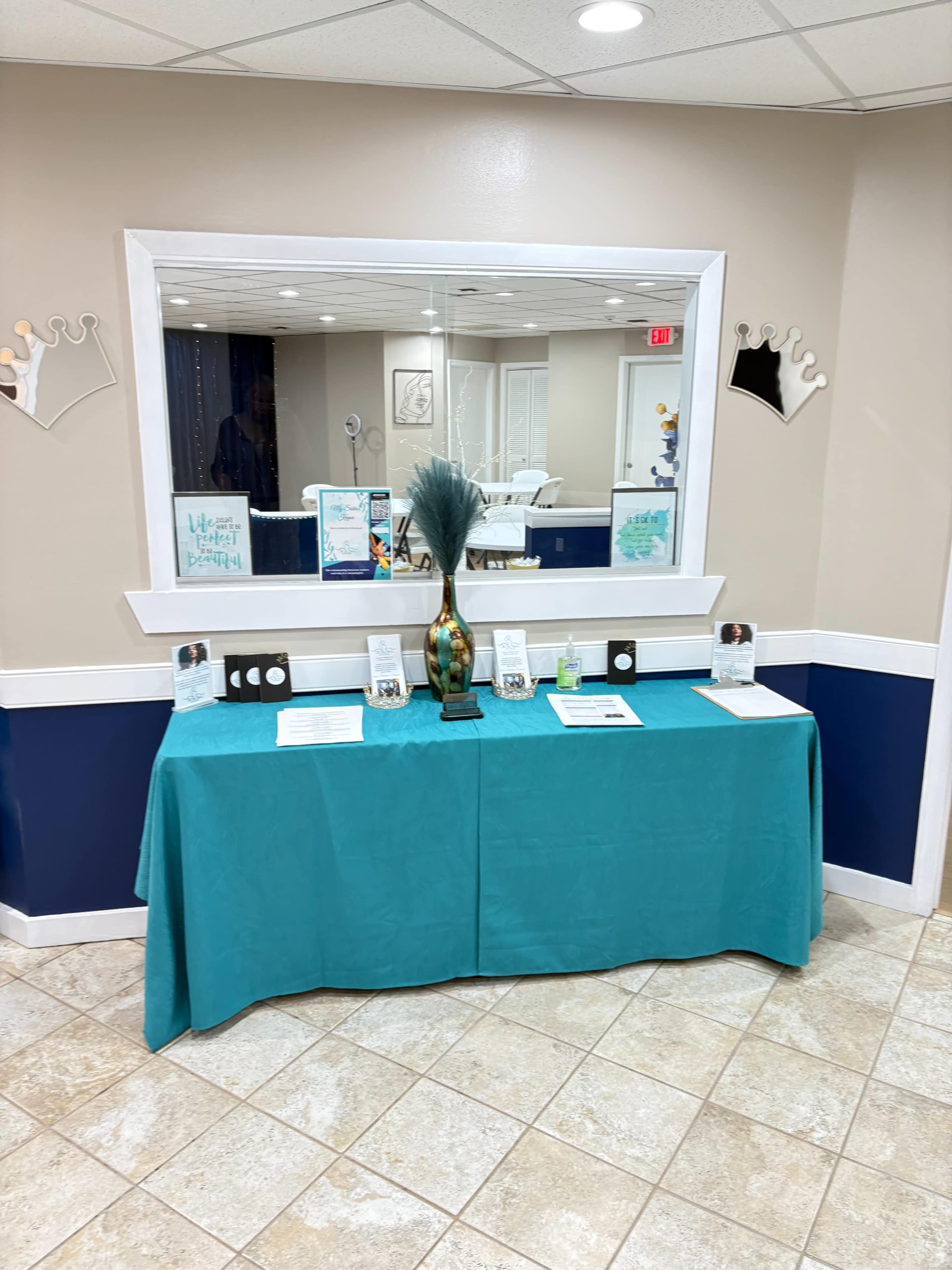 A teal table with informational displays and a decorative vase is positioned against a wall with a large mirror reflecting a waiting area.
