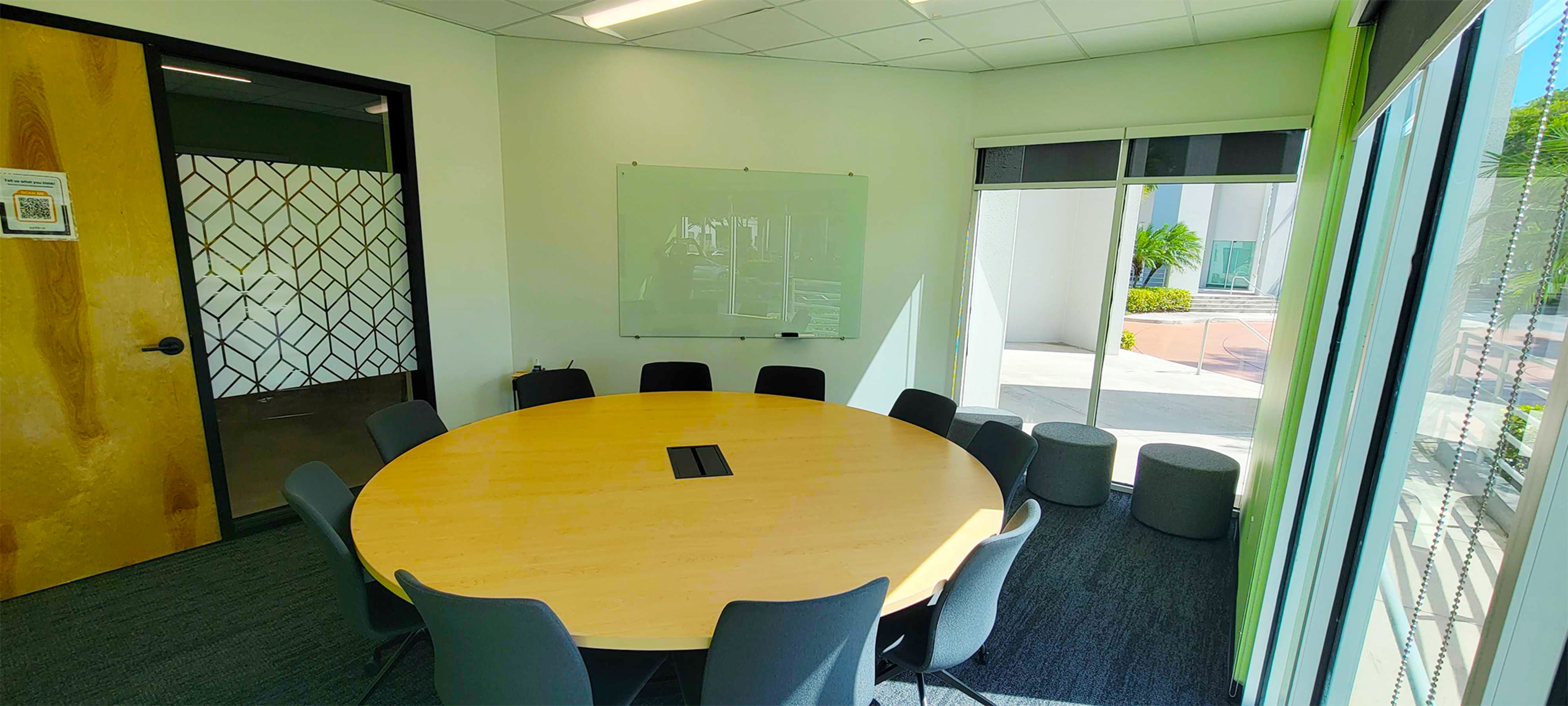 A modern conference room featuring a large circular wooden table surrounded by gray chairs, with a whiteboard on the wall and natural light coming through large windows.