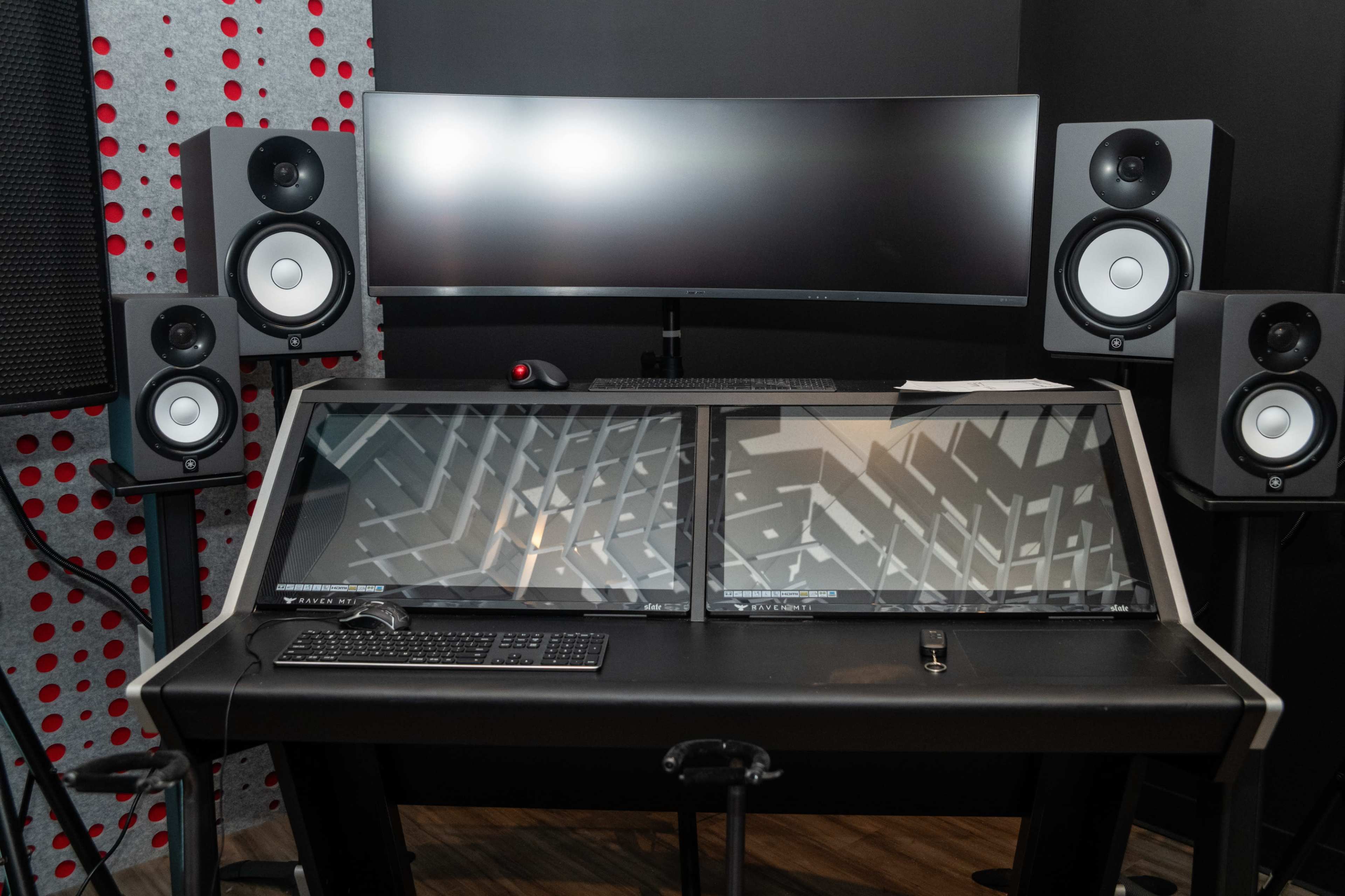 A music production workstation features dual flat screens, a keyboard, and monitors set against a dark backdrop with a patterned wall.