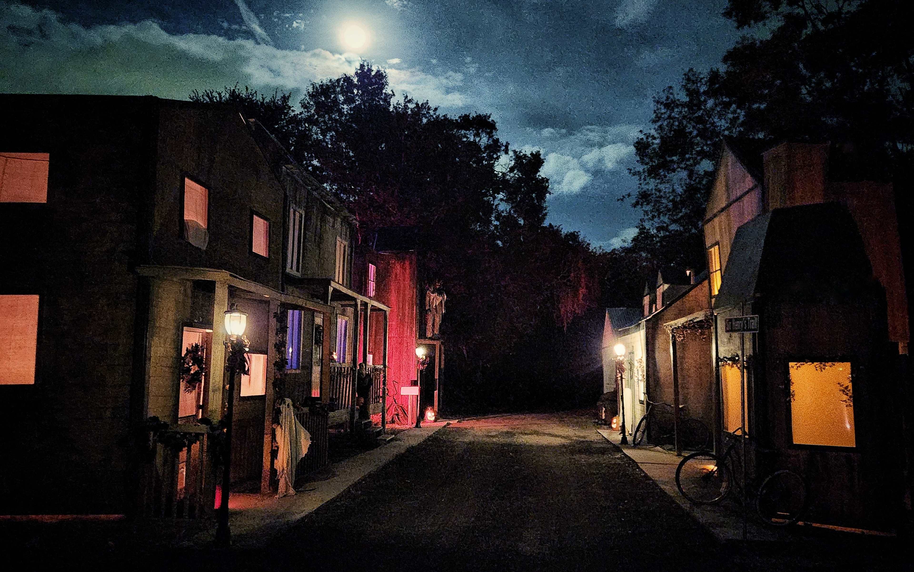 A deserted street scene at night with two rustic buildings on either side and a full moon casting light over the road.