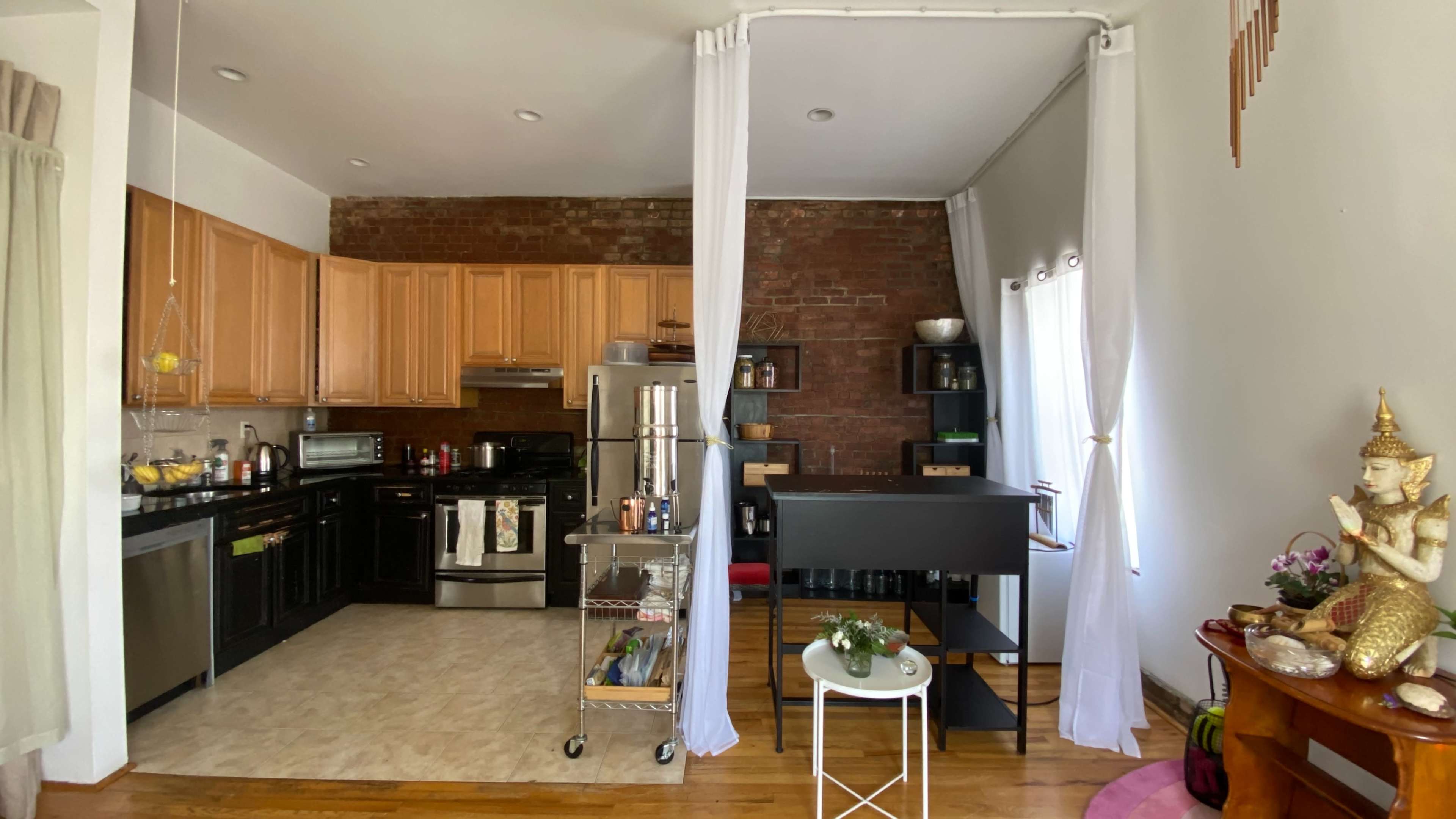 A kitchen with wooden cabinetry and black appliances, separated from a living area by sheer white curtains.