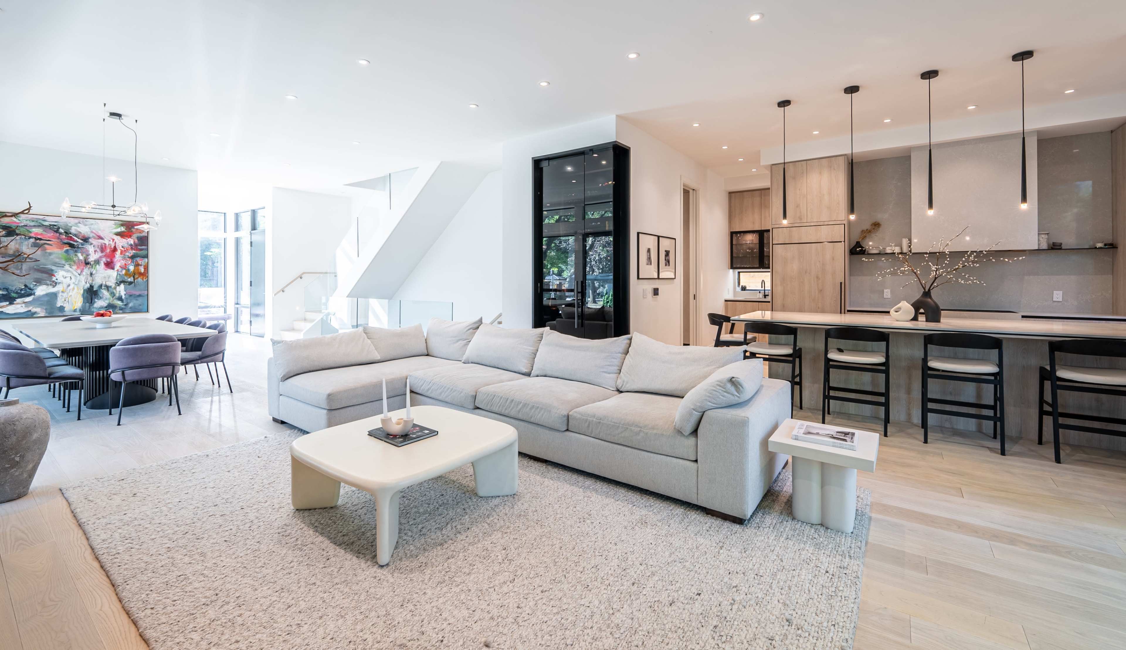 The image depicts a modern, open-concept living space featuring a large sectional sofa, a dining area with a table and chairs, and a sleek kitchen with minimalist cabinetry.