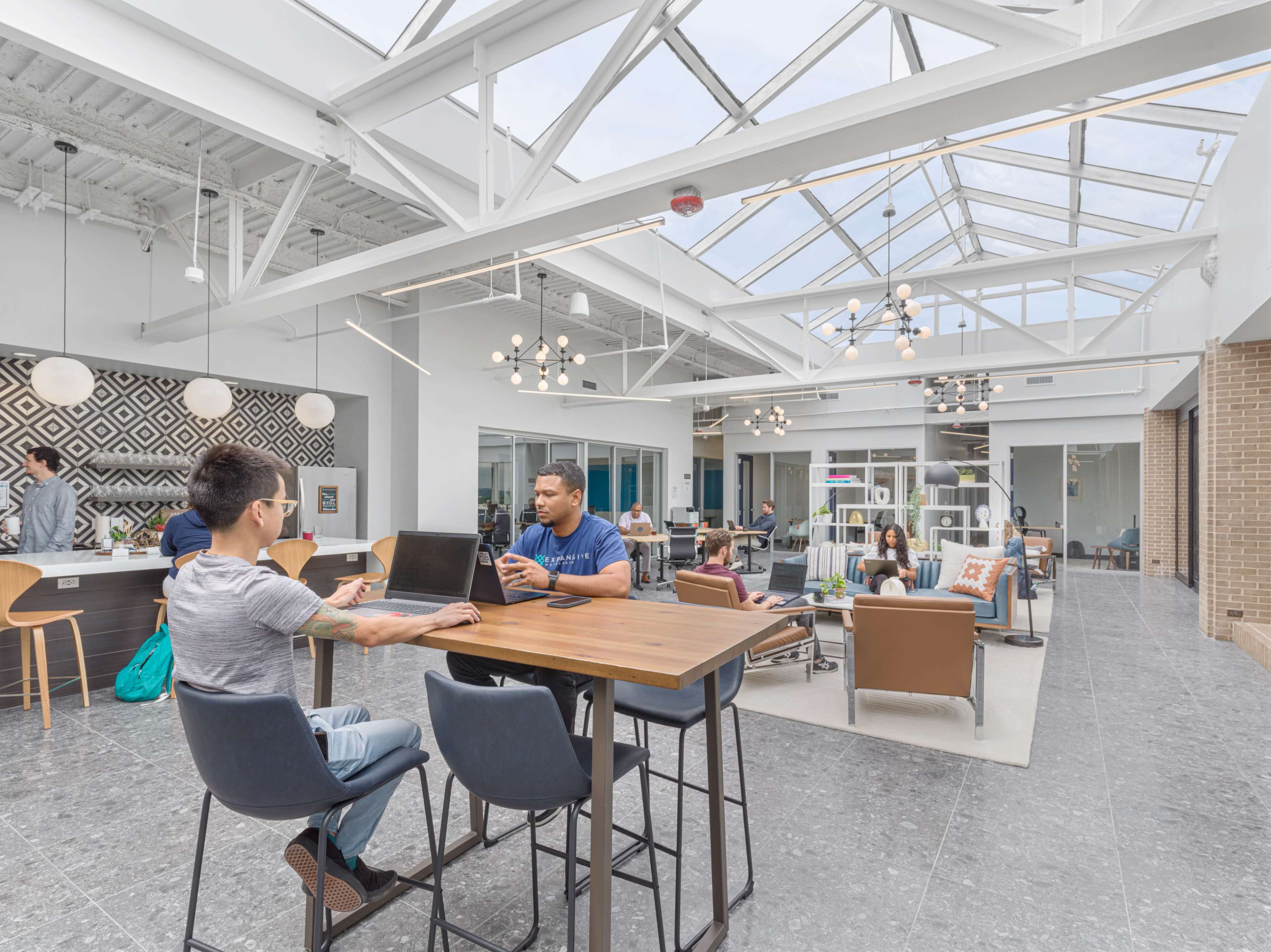 The image shows a modern open-concept workspace with large windows, tables for collaboration, and various seating areas occupied by people engaged in different activities.