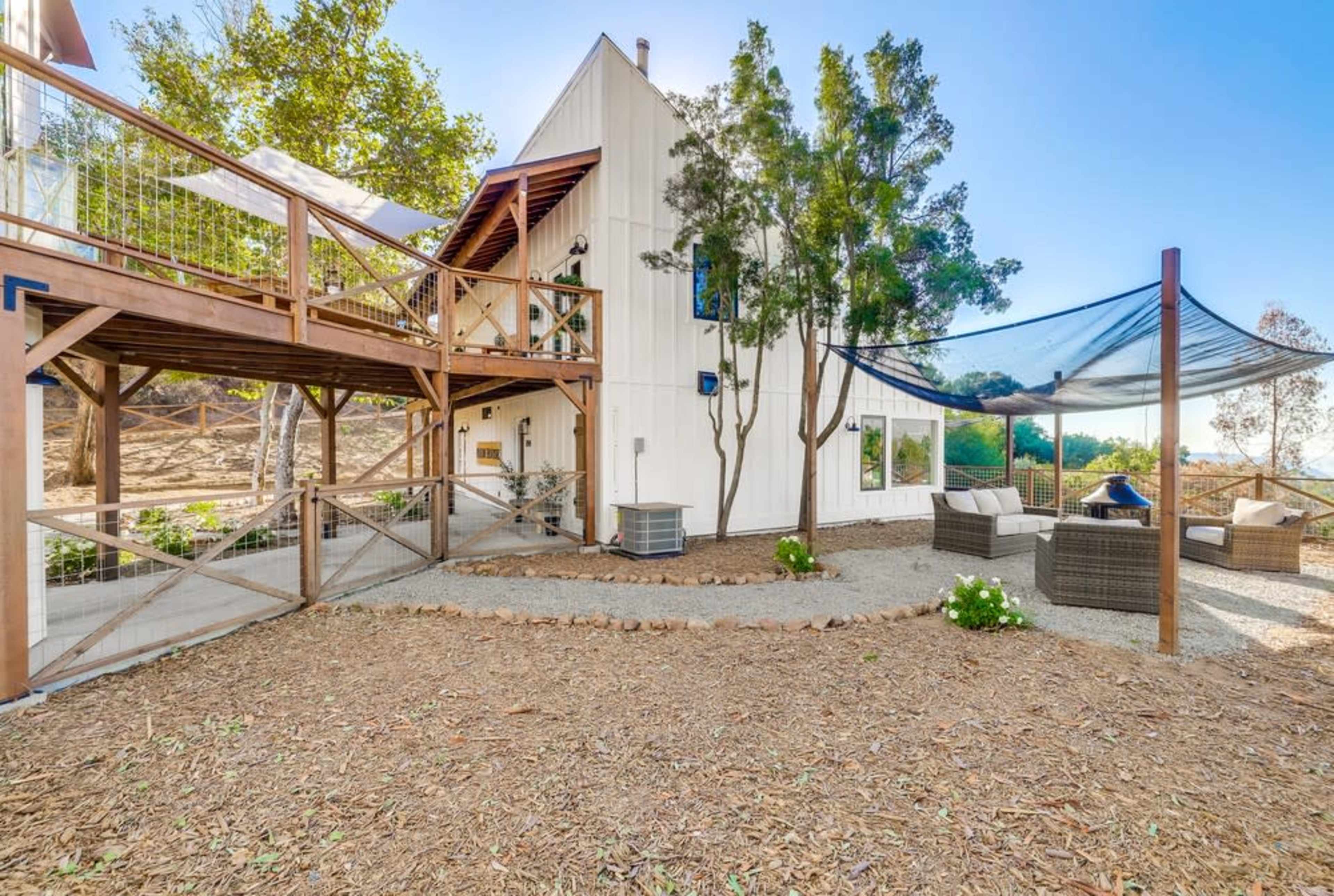 A two-story house with wooden accents features an outdoor seating area surrounded by gravel and trees.