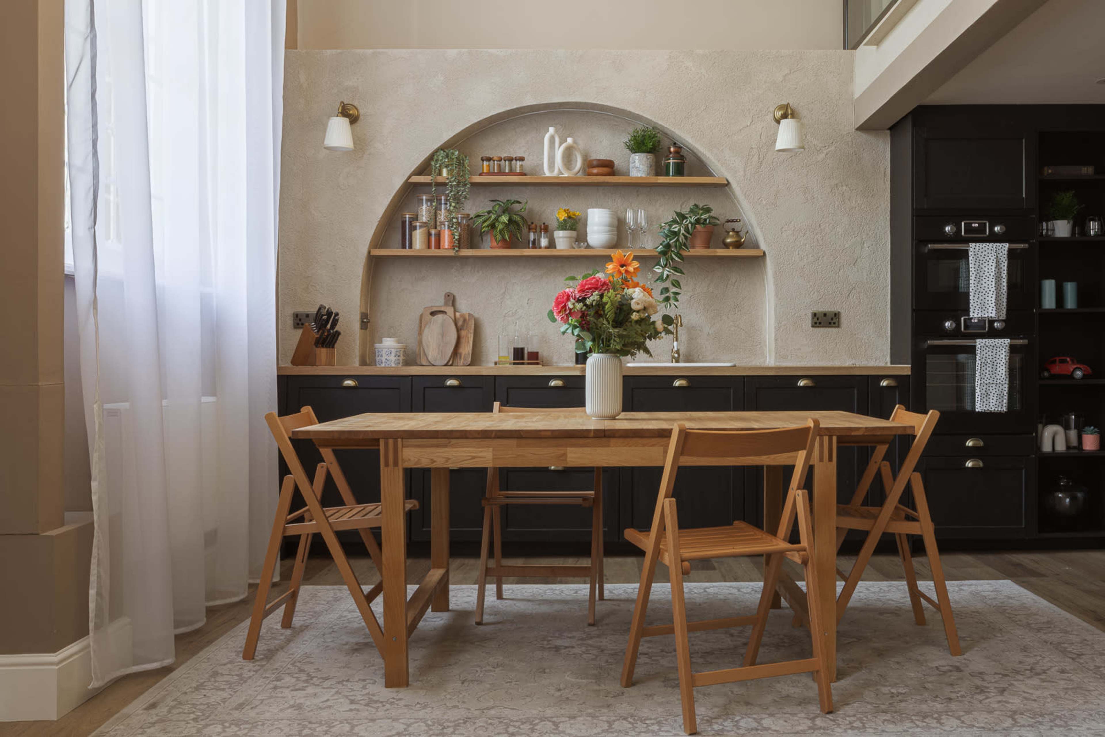 A wooden dining table with four folding chairs is set in a well-lit room featuring a textured wall with a curved shelf displaying plants and kitchen items.