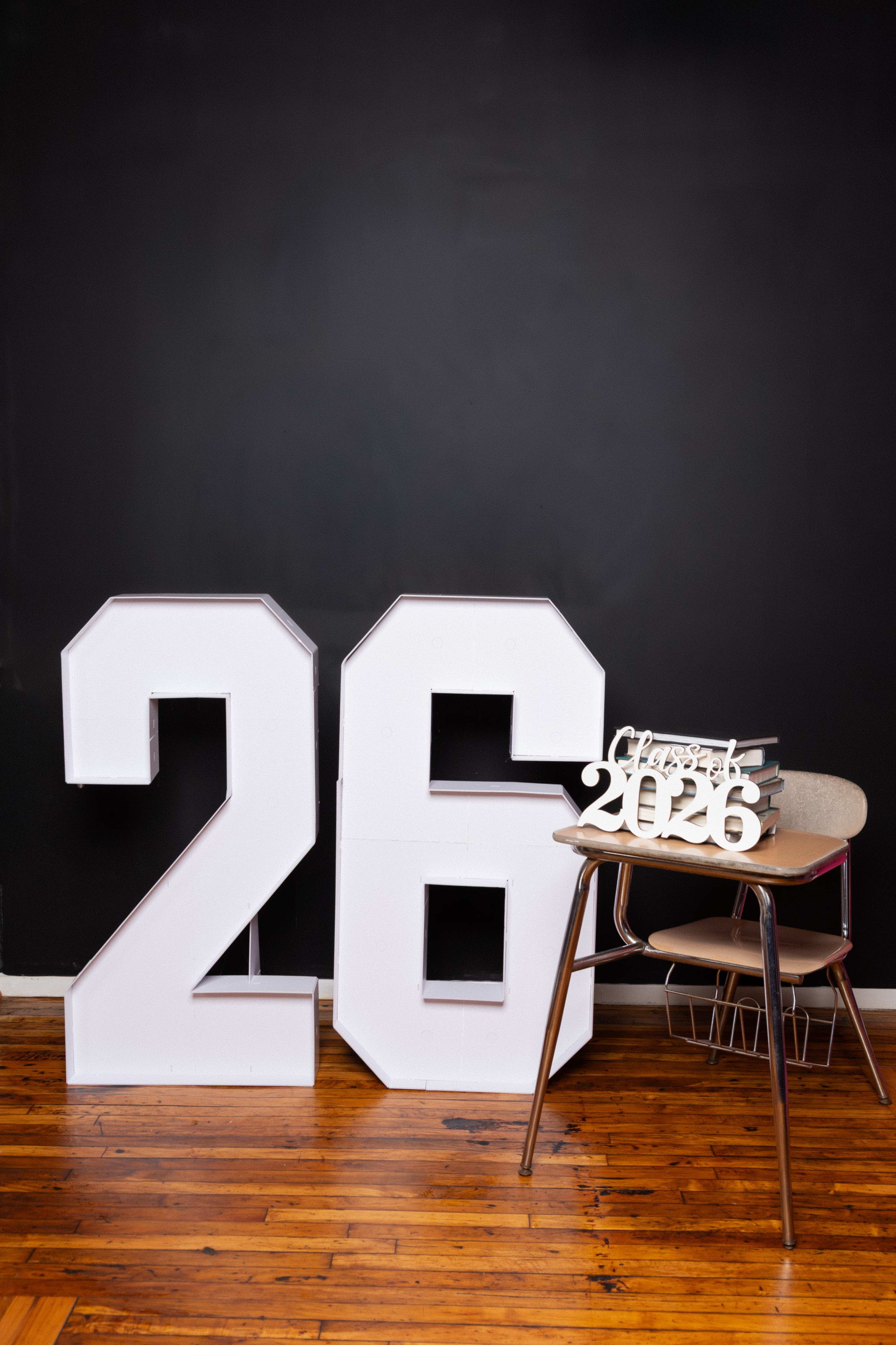 The image features large white numbers "26" against a black wall, alongside a small chair and a decorative item reading "Class of 2026" on a wooden floor.