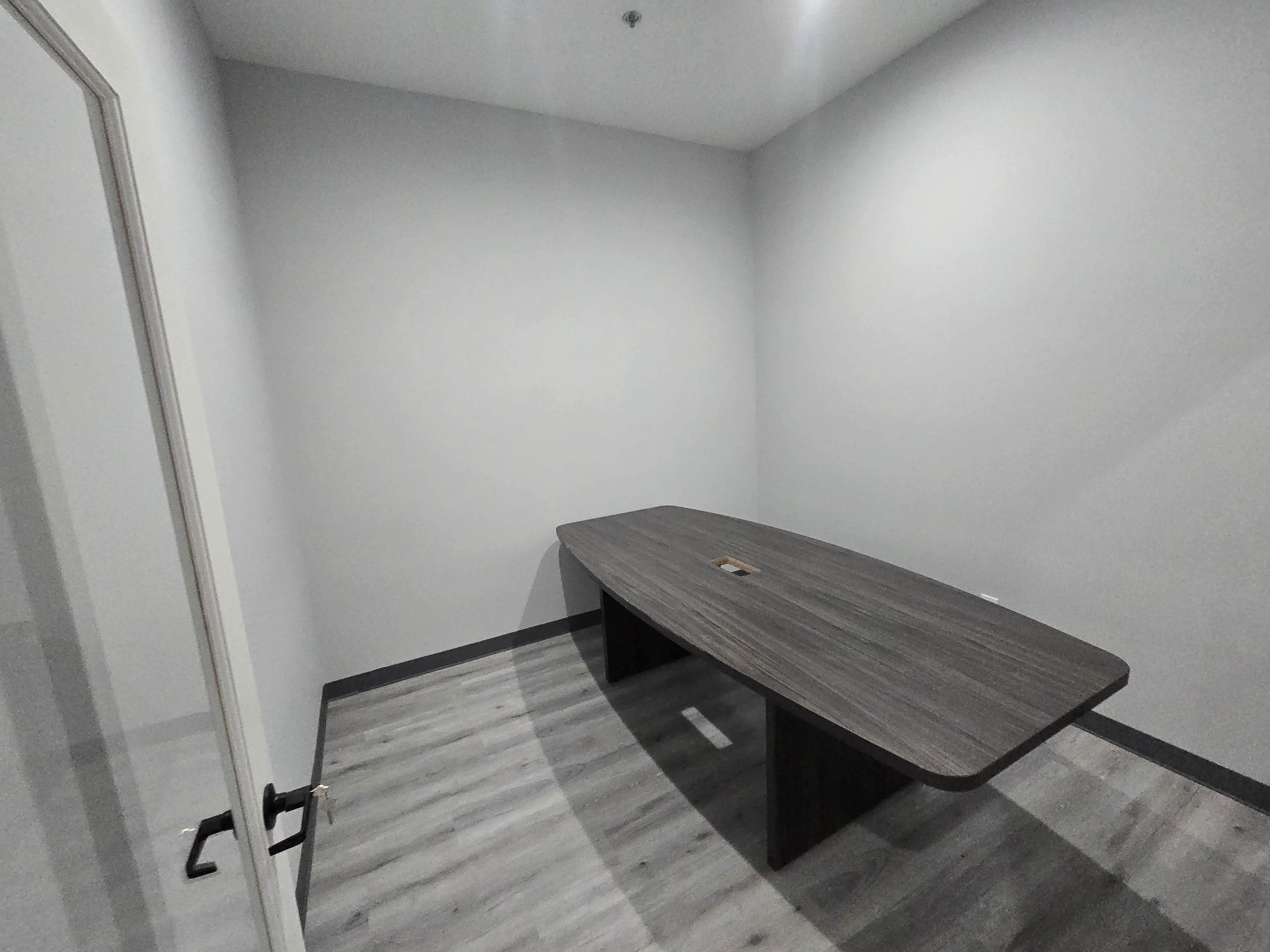 The image shows a sparsely furnished meeting room with a large, rectangular table and gray walls.