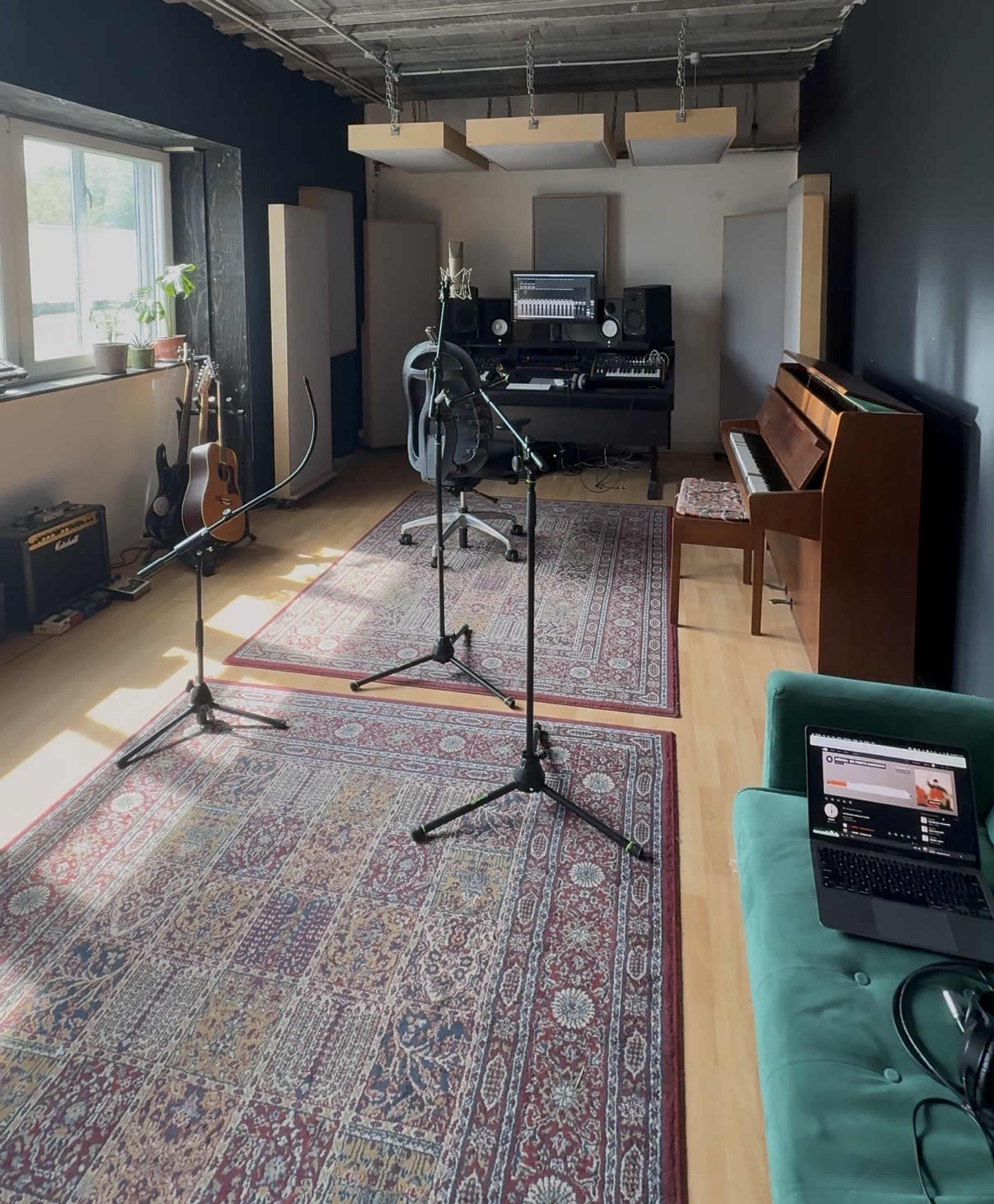 The image shows a music recording studio featuring a grand piano, microphones on stands, an audio mixer setup, and various instruments on display in a spacious, well-lit room.