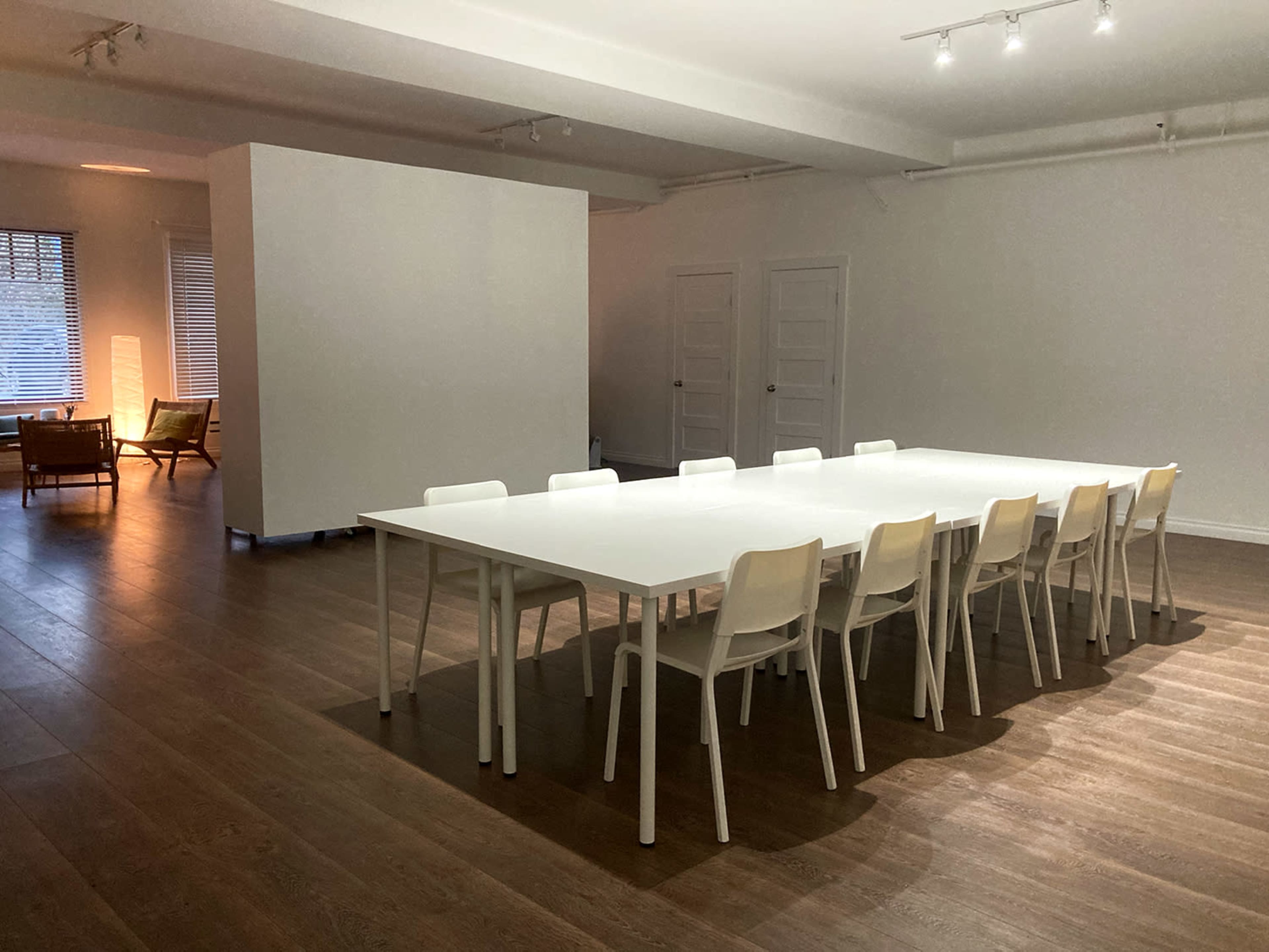 A large, empty meeting room features a long white table surrounded by ten white chairs, with wooden flooring and a partition in the background.