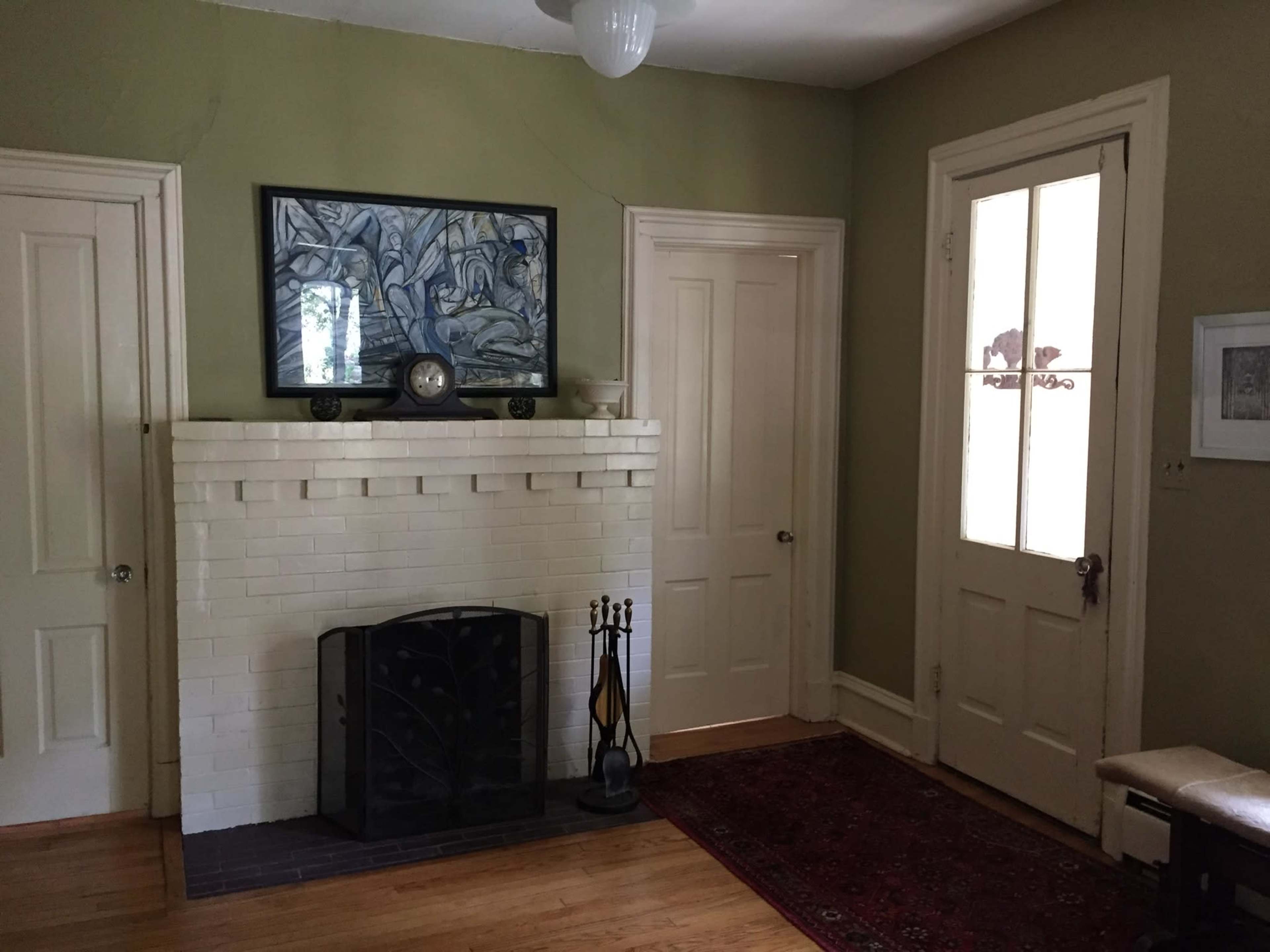 The image shows a simple entryway with a white brick fireplace, framed artwork on the wall, a wooden door, and a small area rug.