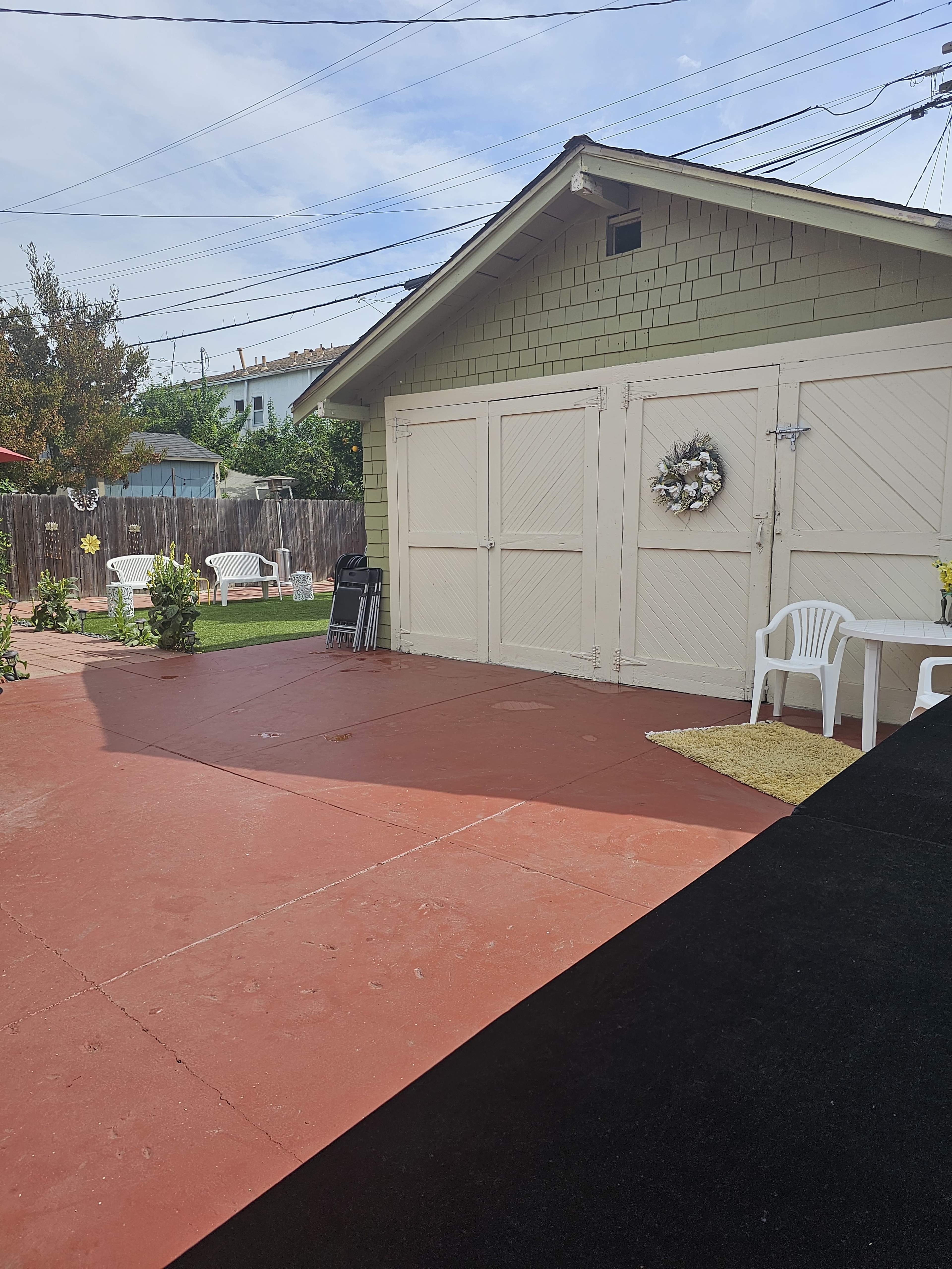 The image shows a backyard with a red concrete patio, a garden shed, and white patio chairs arranged on the left.