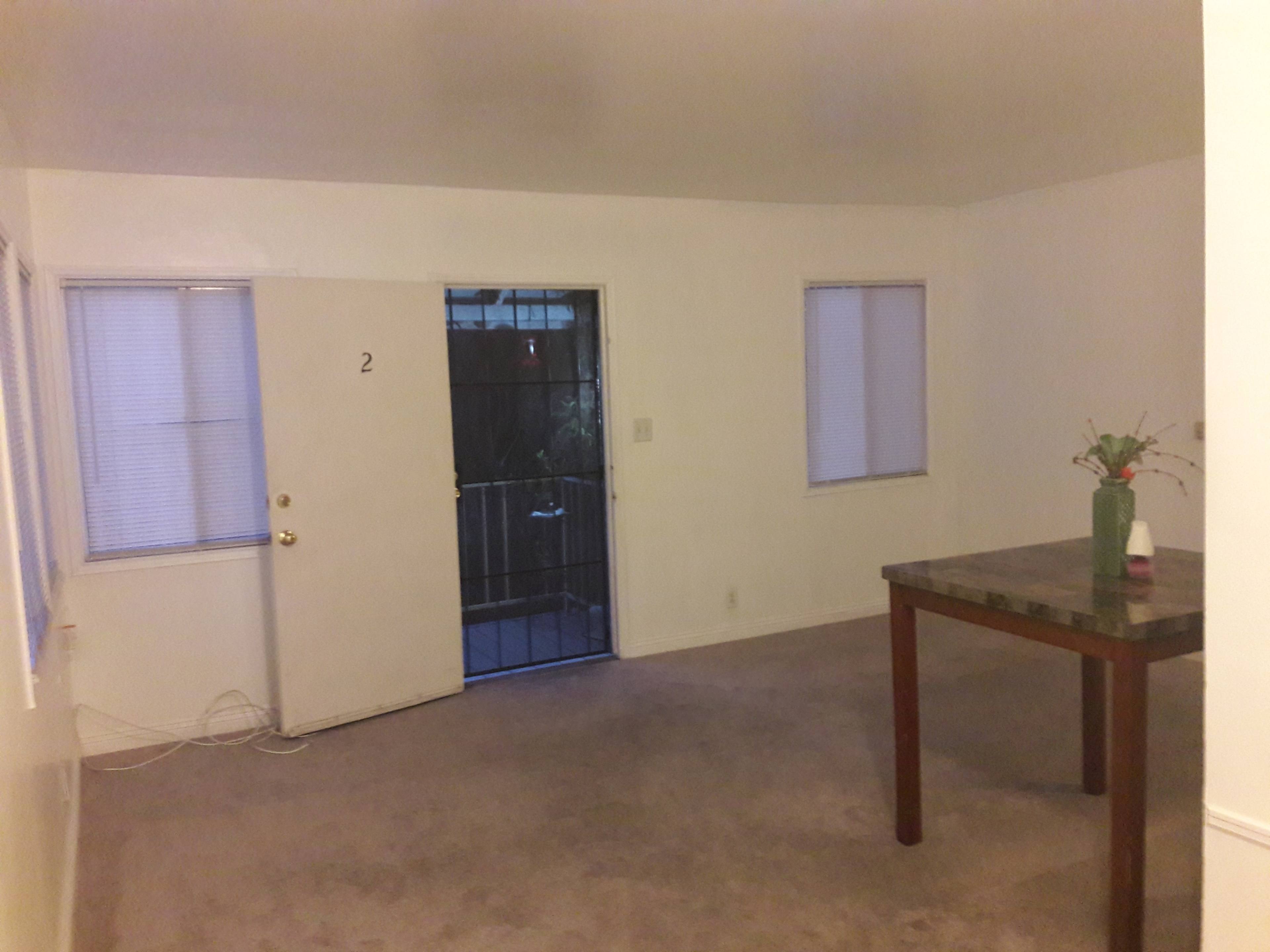 The image shows a sparsely furnished room with a door leading outside, a table in the center, and two windows on adjacent walls.
