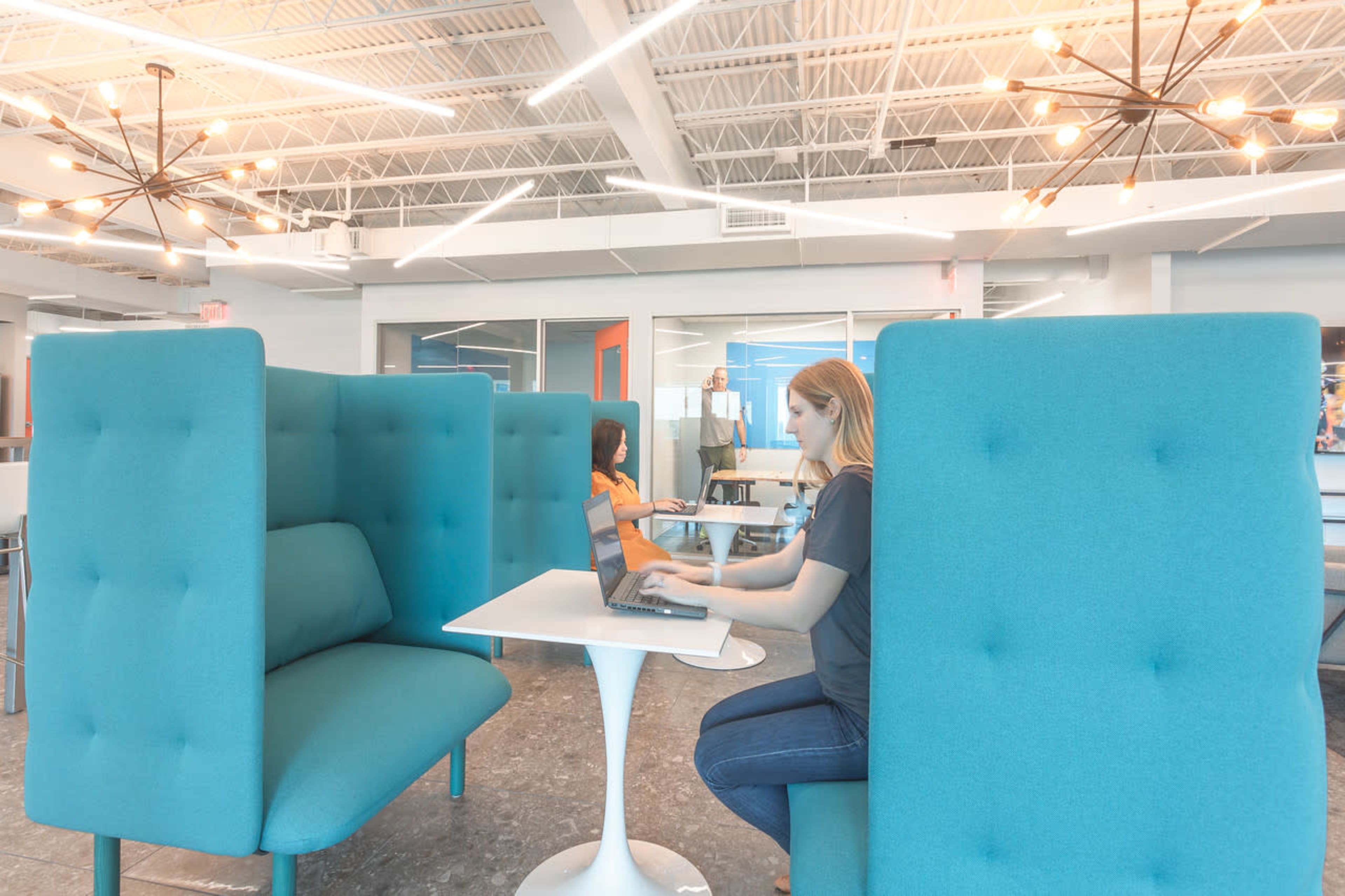The image shows two people working in a modern office space with blue acoustic booths and pendant lighting.