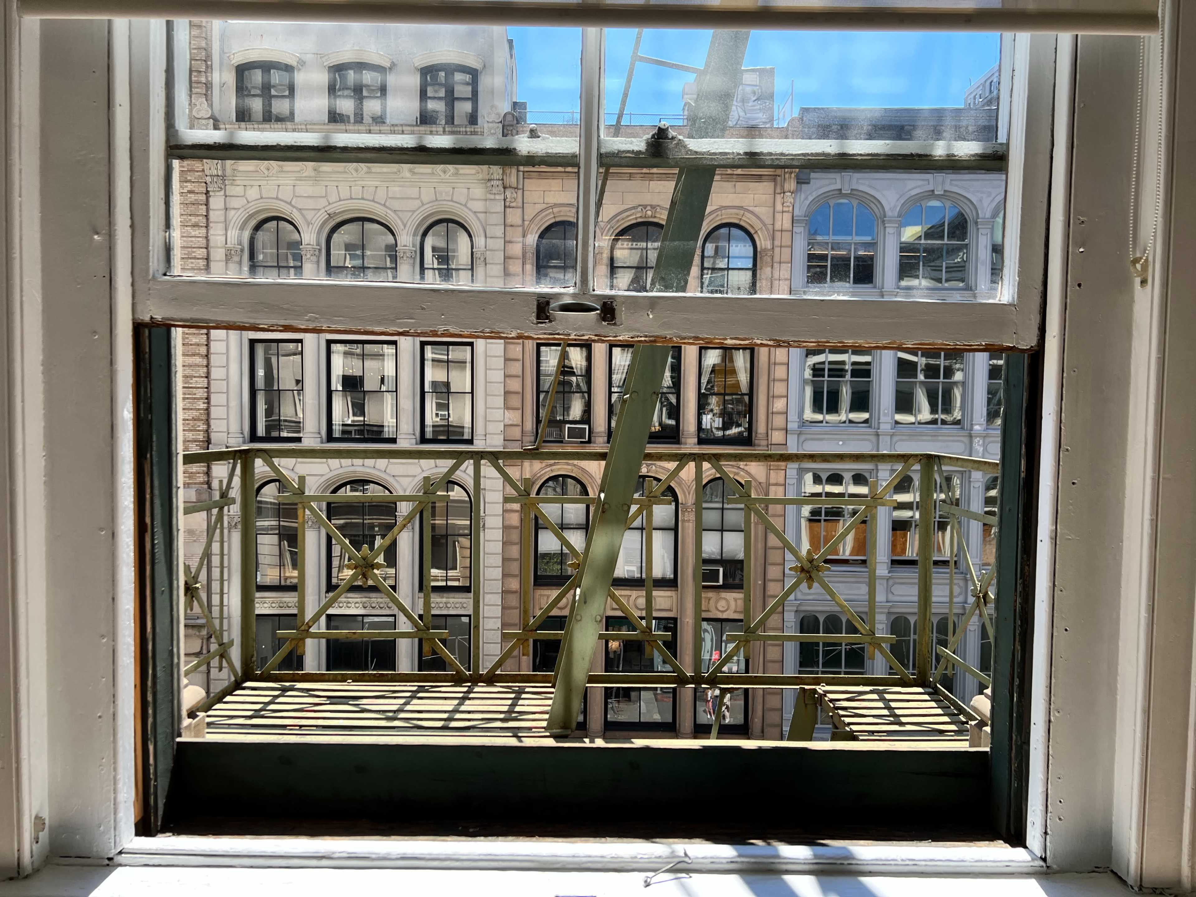 The image shows a view through a window featuring a green balcony railing with surrounding buildings and blue sky in the background.