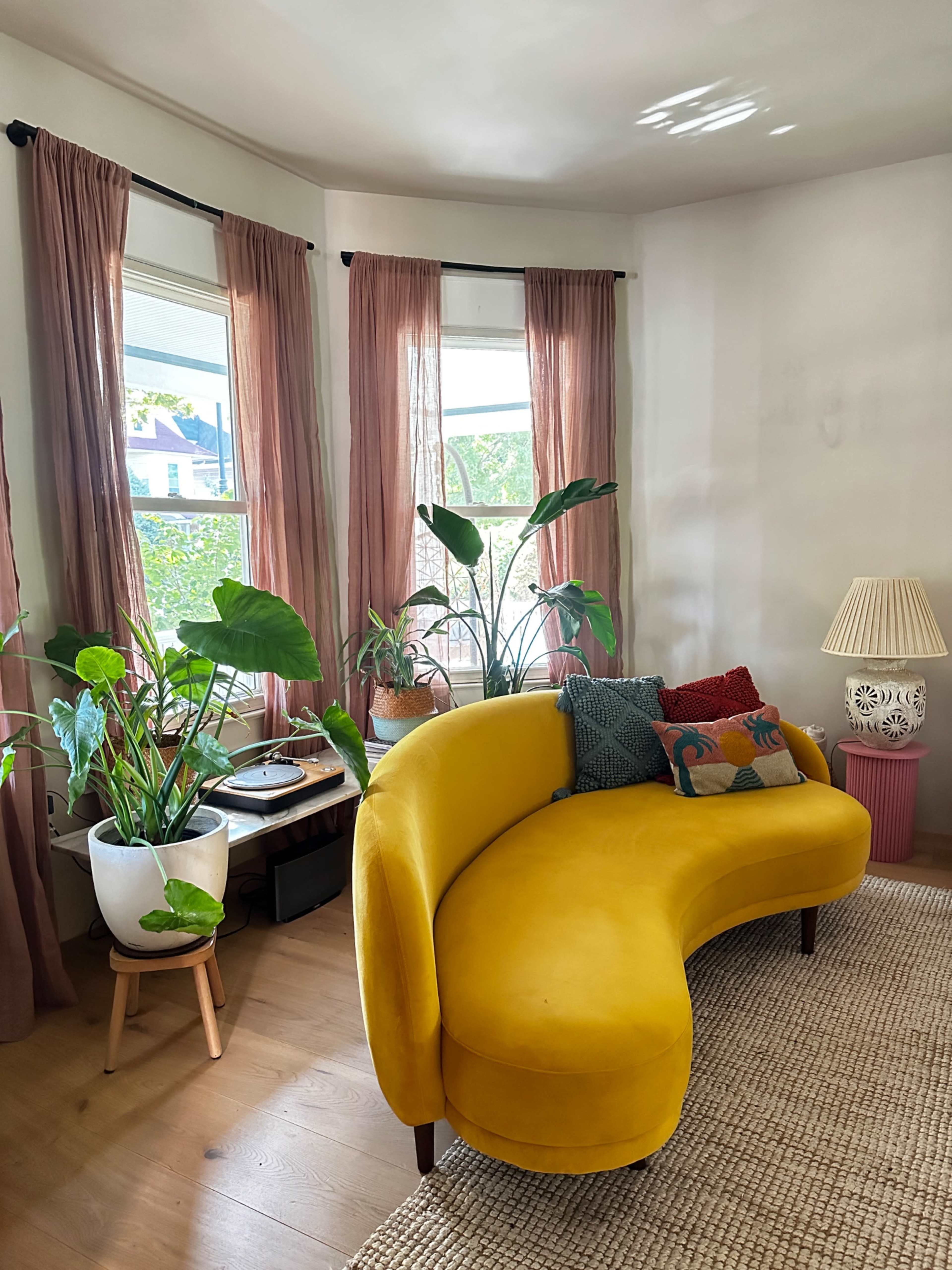 A curved yellow couch is positioned in a sunlit room with large windows draped in pink curtains, surrounded by various indoor plants and decorative cushions.