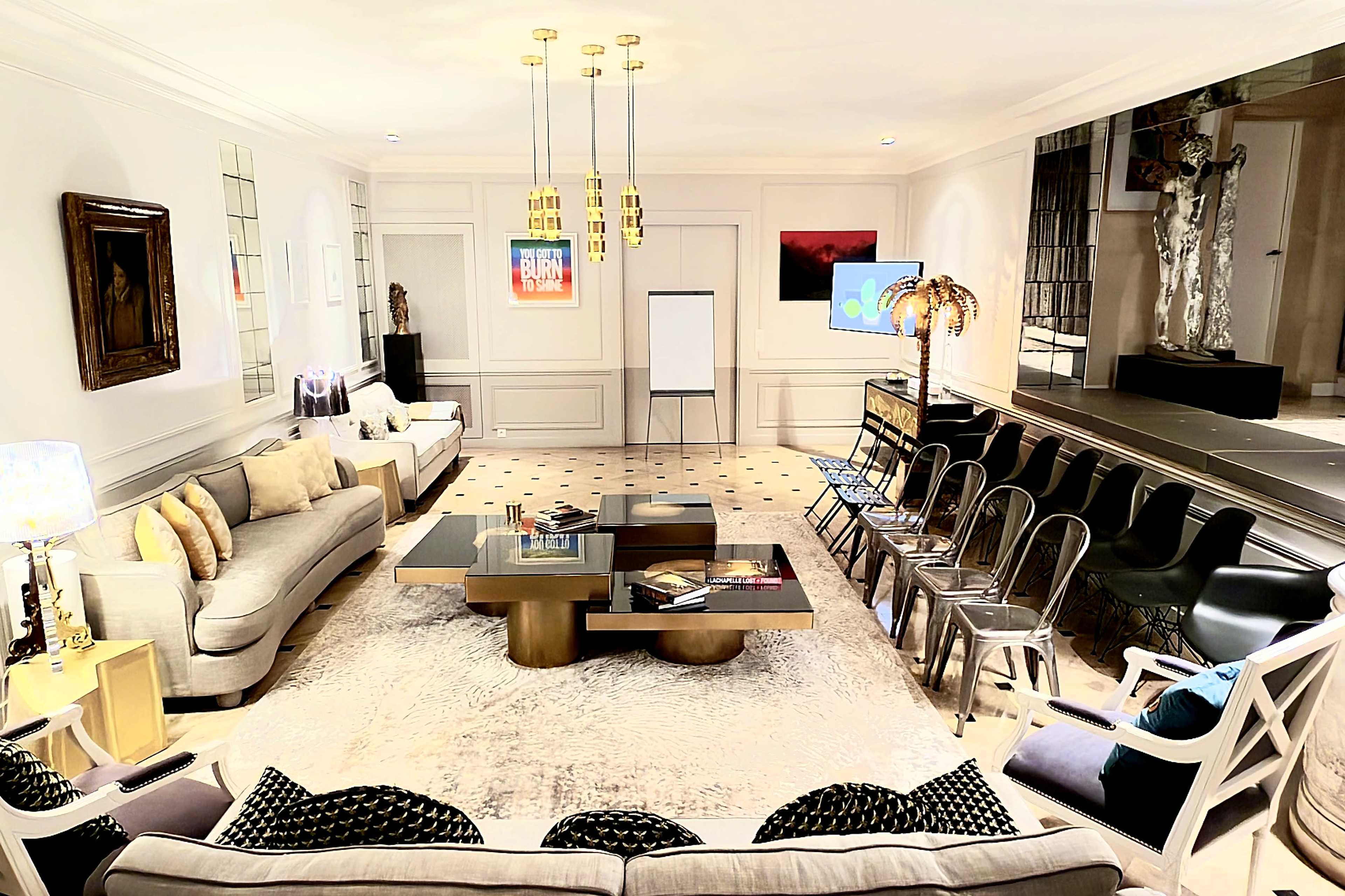 The image shows a modern meeting space with a large seating area, a central coffee table, and several chairs arranged for a presentation or discussion, illuminated by stylish lighting fixtures.