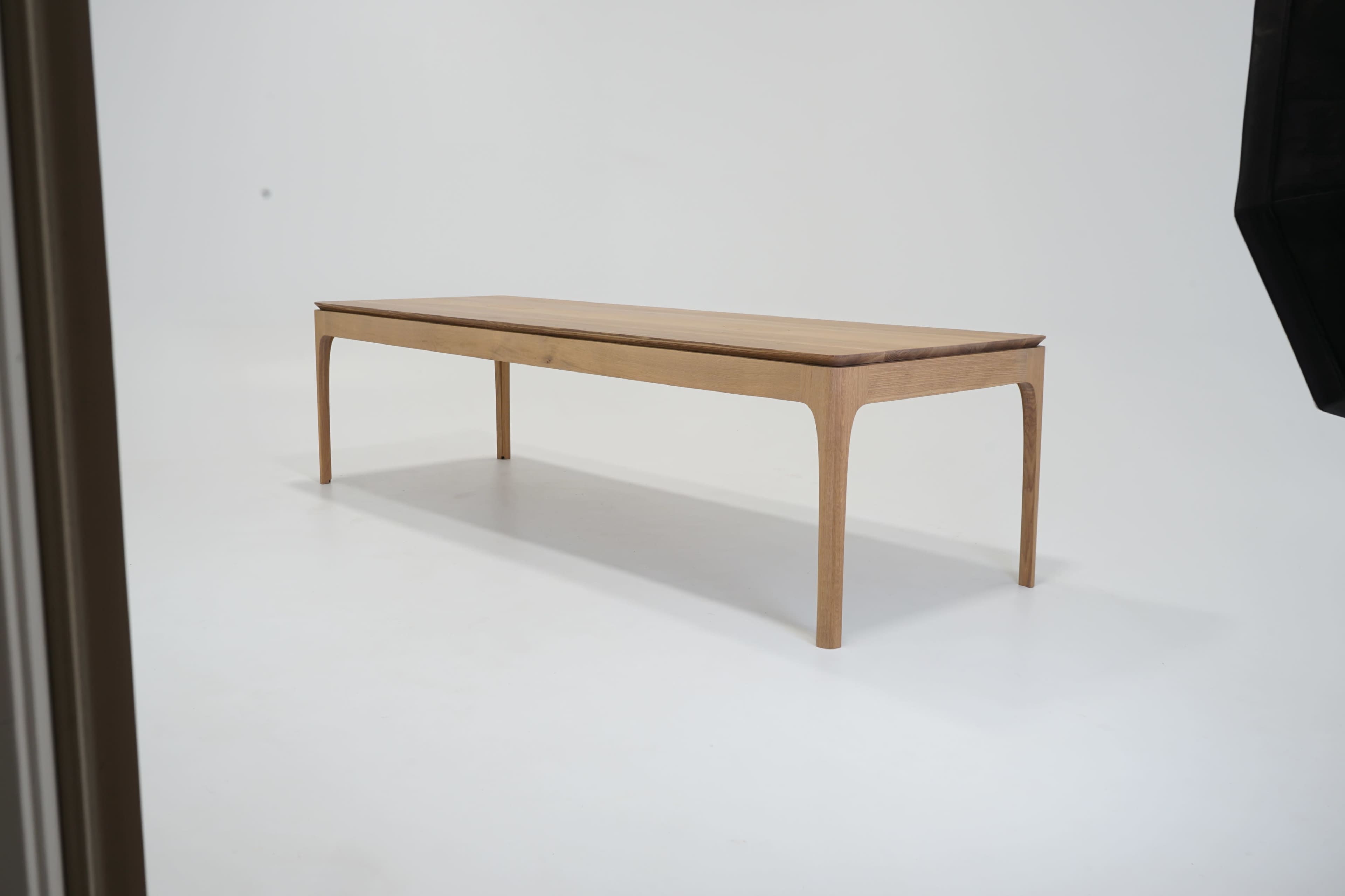 A wooden table with slender legs is positioned in a well-lit, minimalist studio setting with a white background.