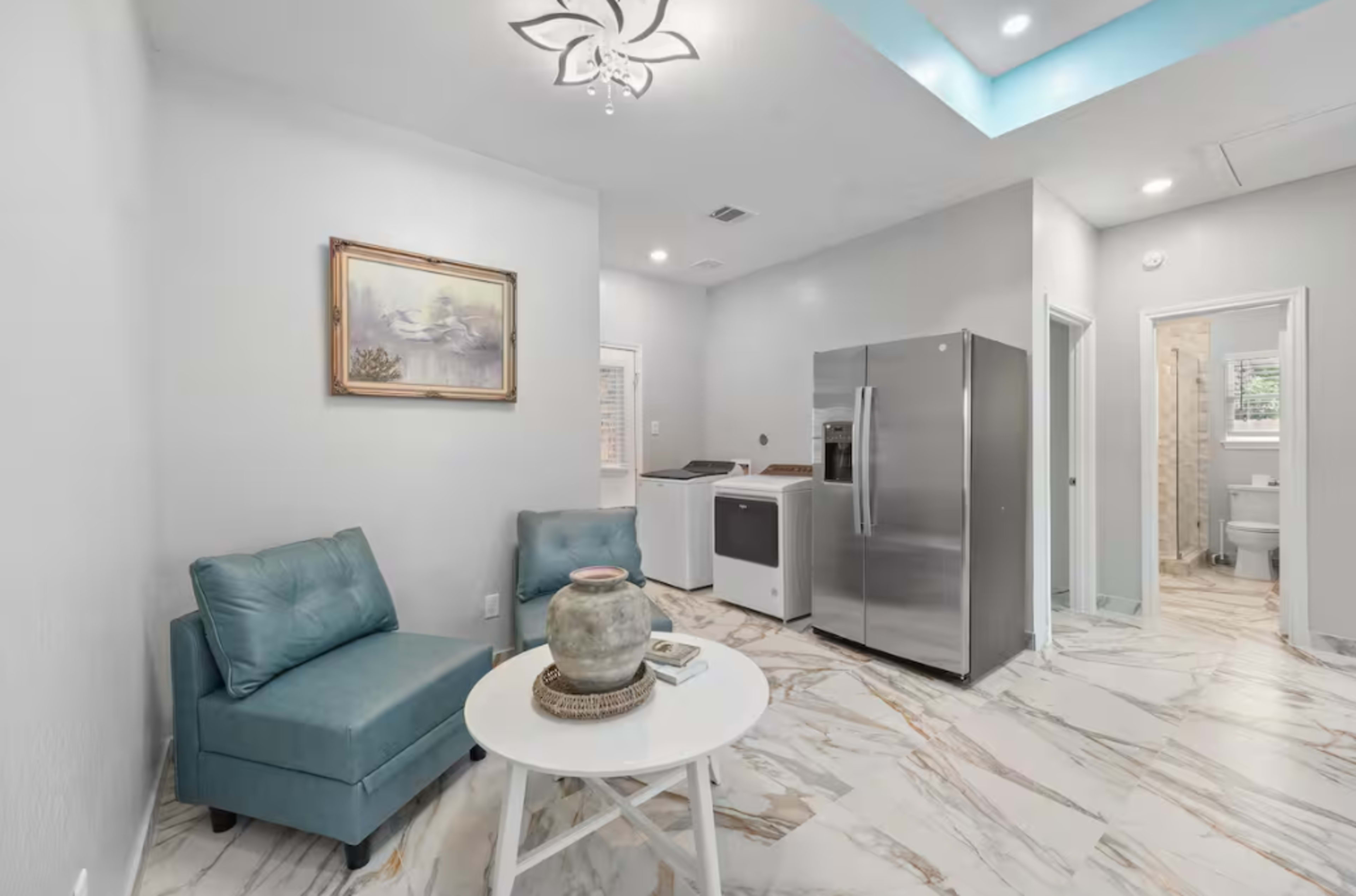 The image shows a modern living space featuring a light-colored tile floor, a small round table with a decorative vase, a teal armchair, and a stainless steel refrigerator.