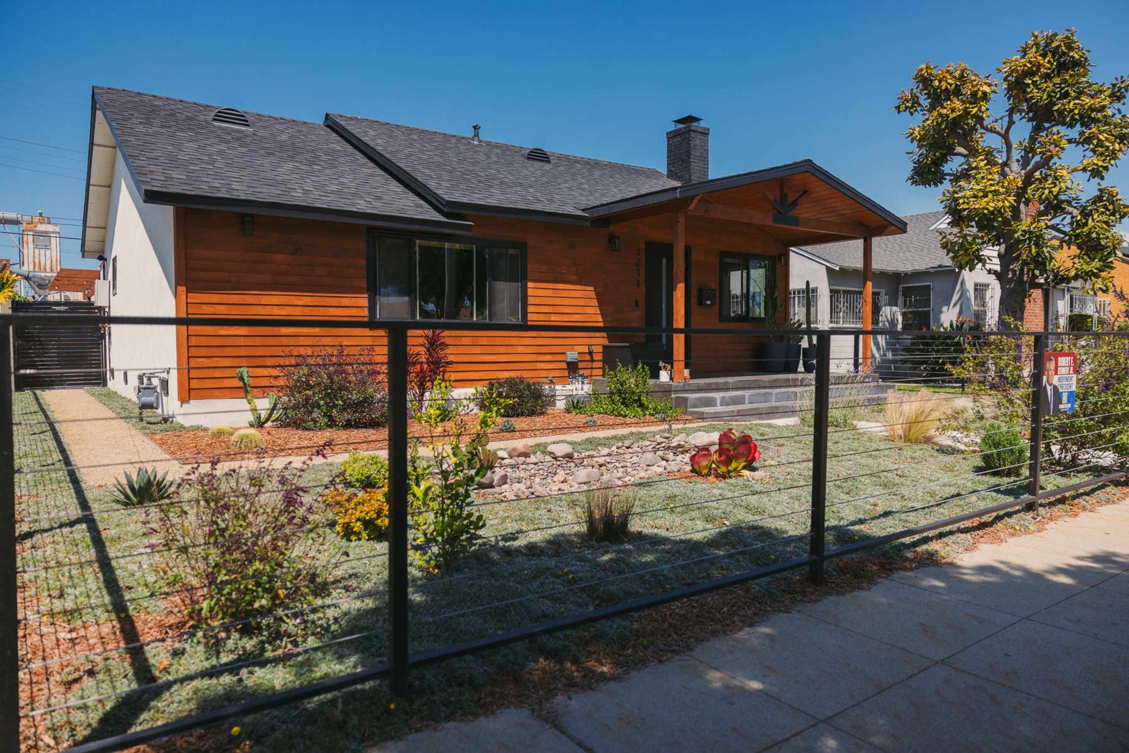 A modern single-story house with a wooden facade and a landscaped front yard is separated from the sidewalk by a black metal fence.