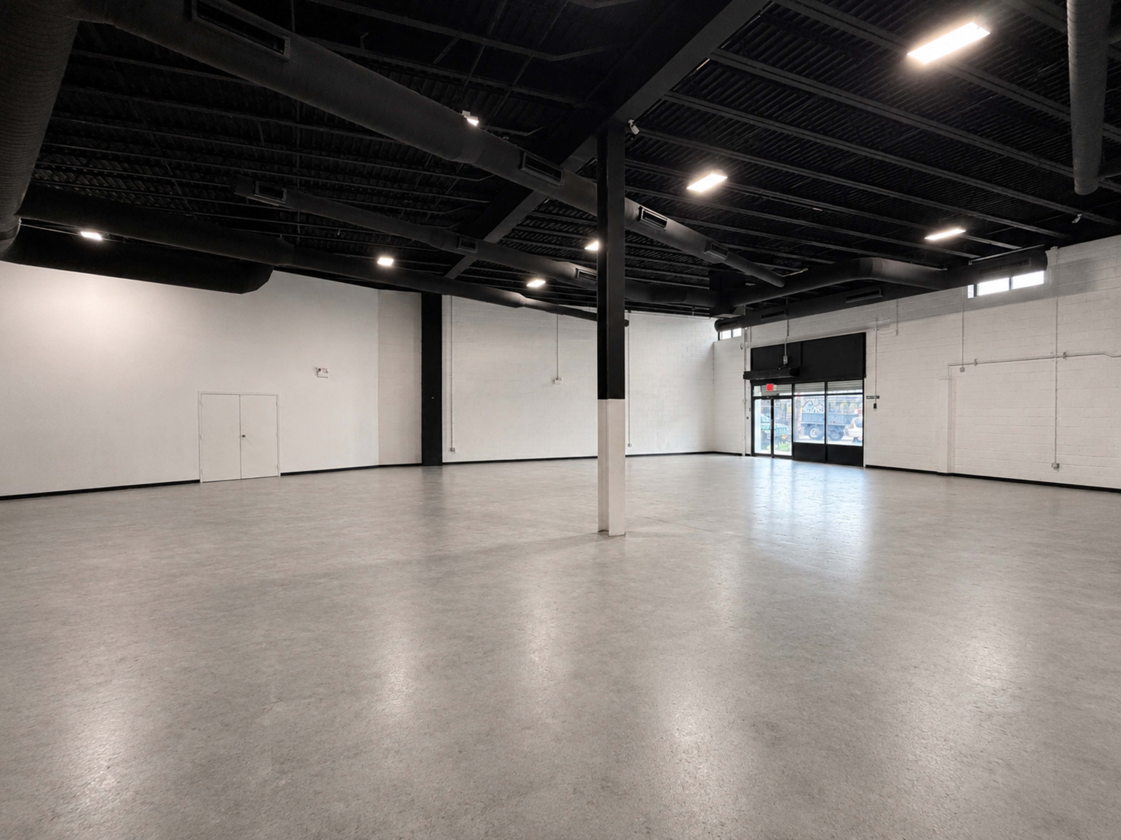 The image shows a spacious, empty indoor area with gray flooring, black ceiling beams, and large windows along one wall.