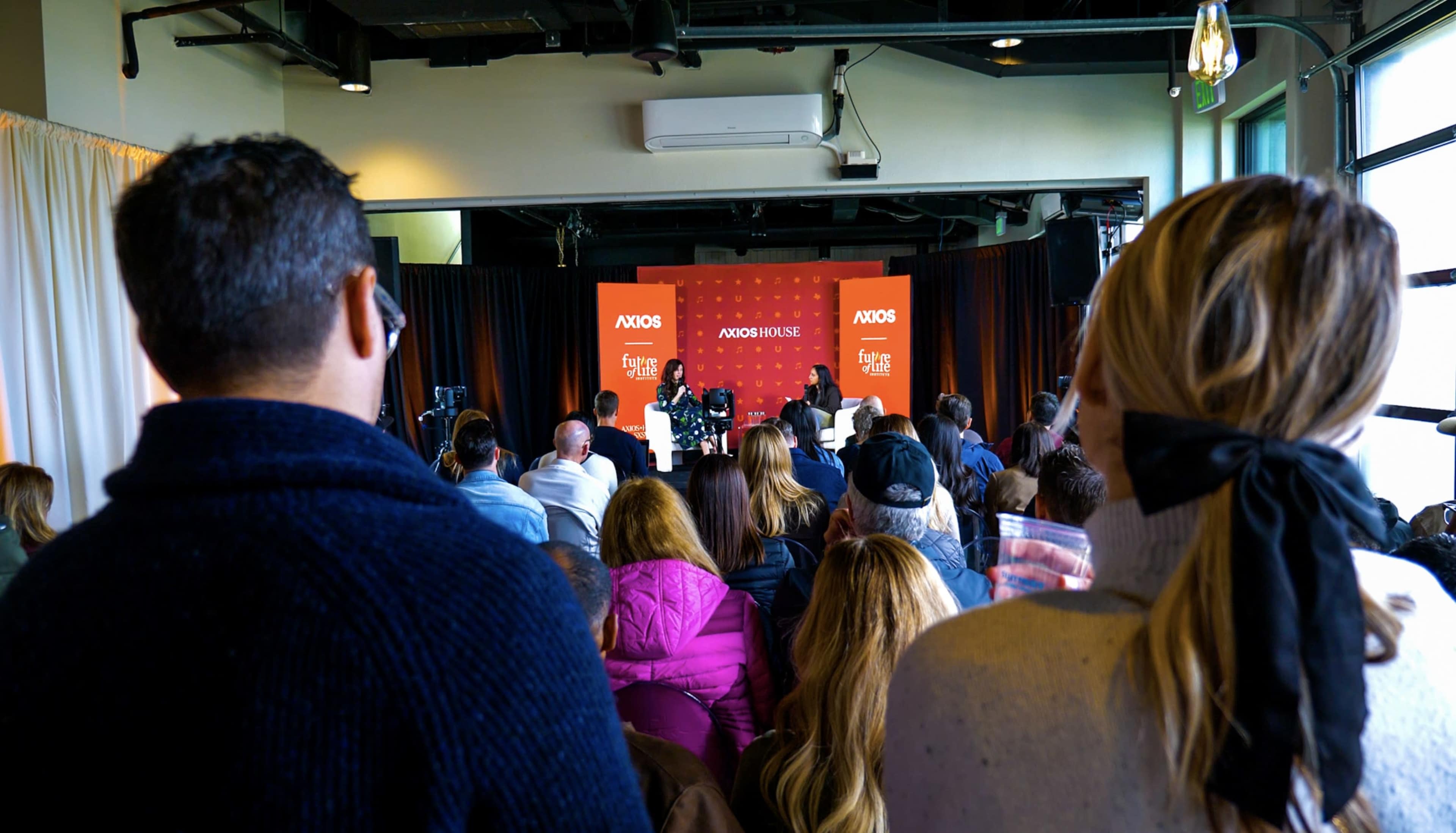 A crowd attends an event featuring speakers on a stage in front of a backdrop with the Axios logo.