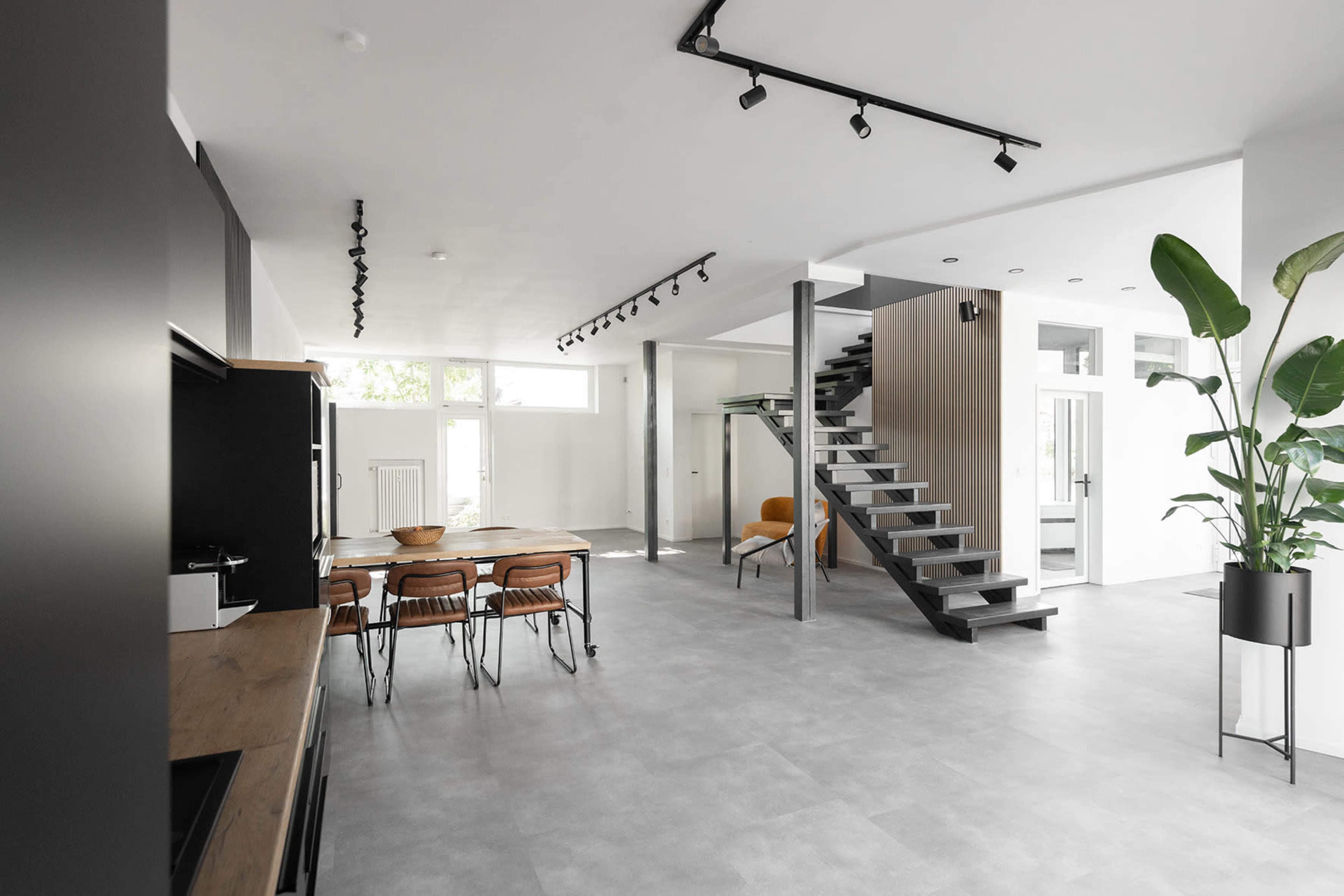 A modern, open-concept living space features a kitchen area with a dining table, a staircase leading to an upper level, and large windows allowing natural light to enter.