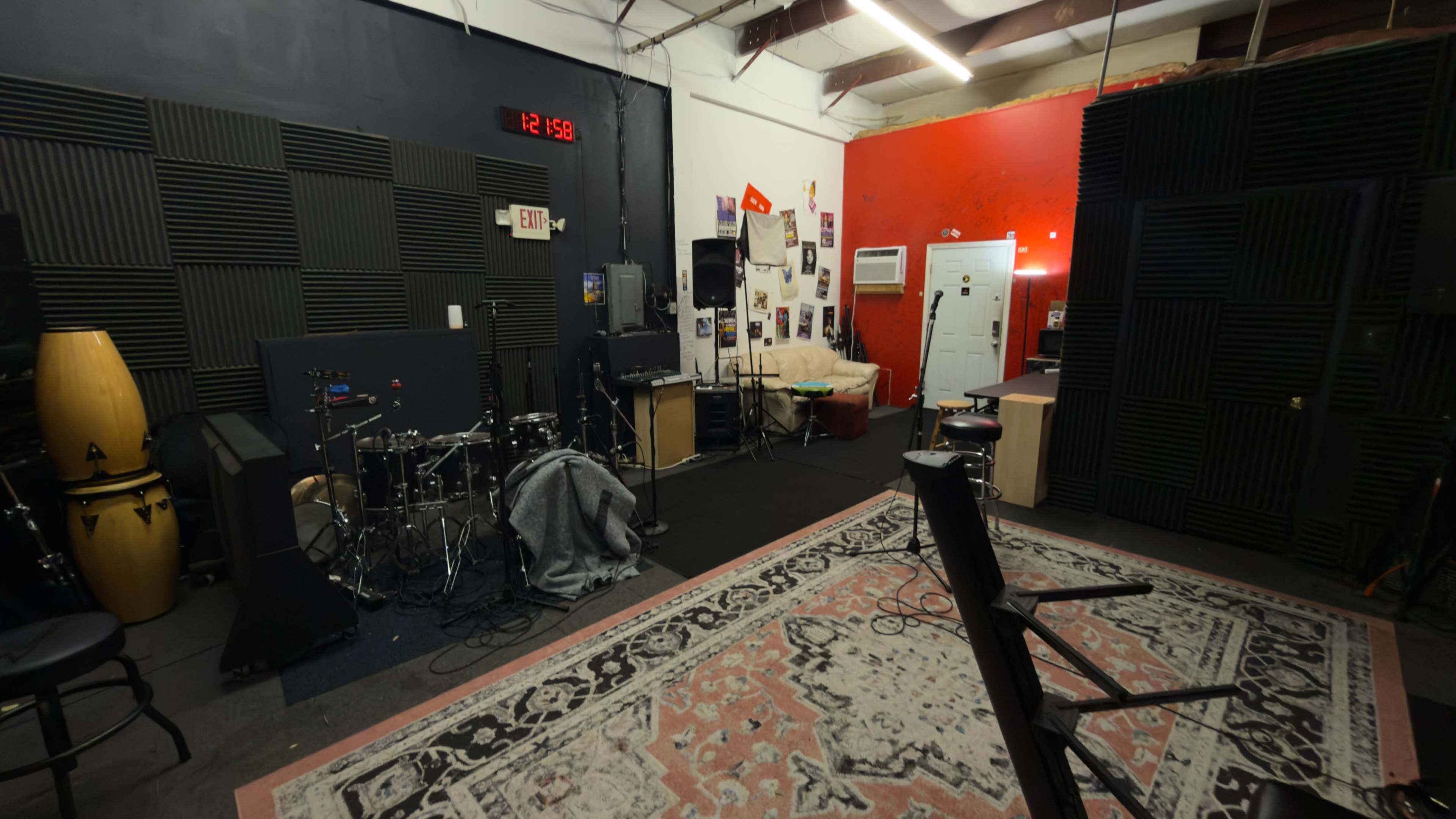 The image shows a music rehearsal space with a drum set, amplifiers, and a large area rug, surrounded by soundproofing panels.