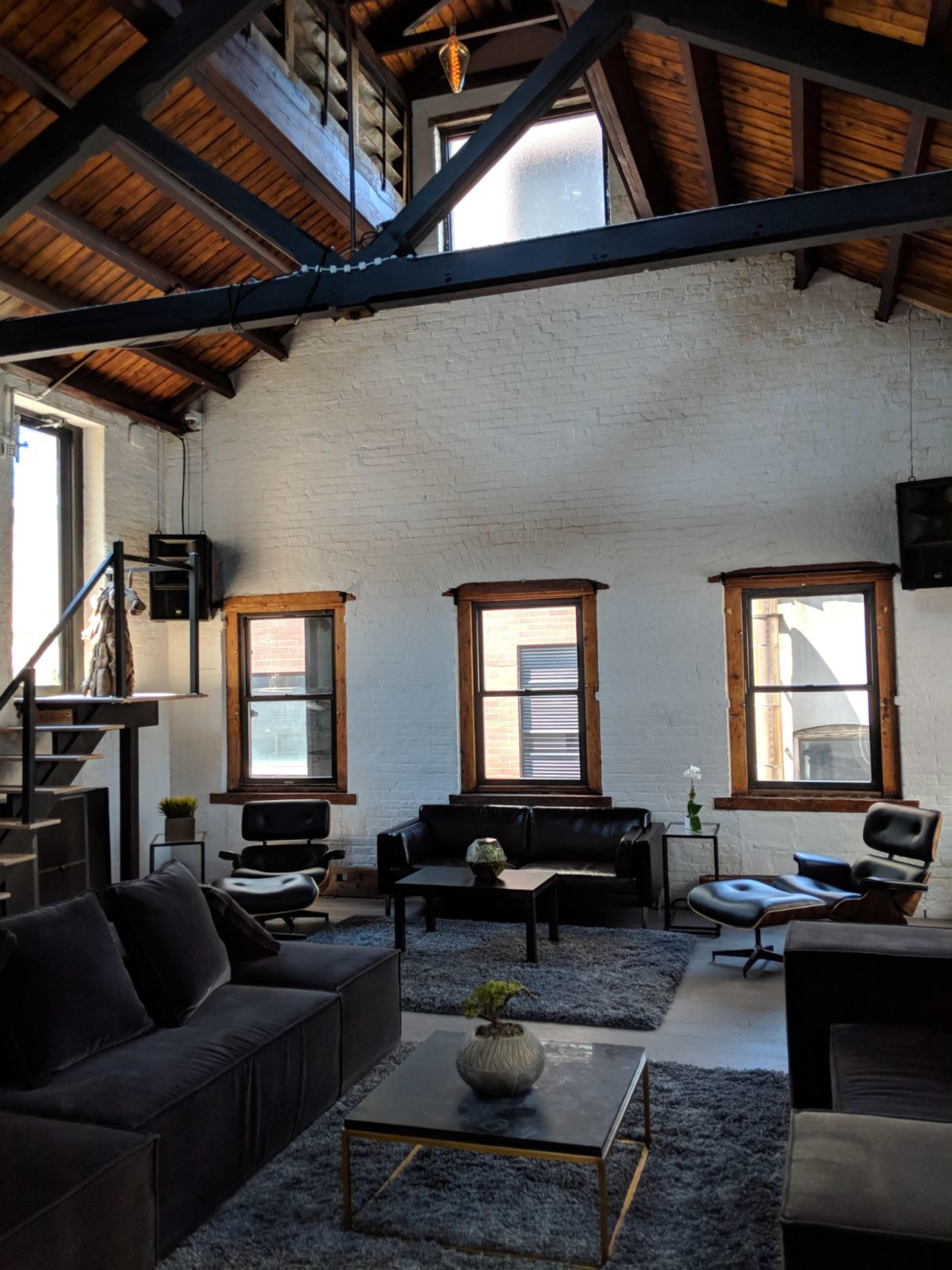 A spacious, modern living area with high ceilings, exposed beams, large windows, and minimalist furniture arranged around a coffee table.