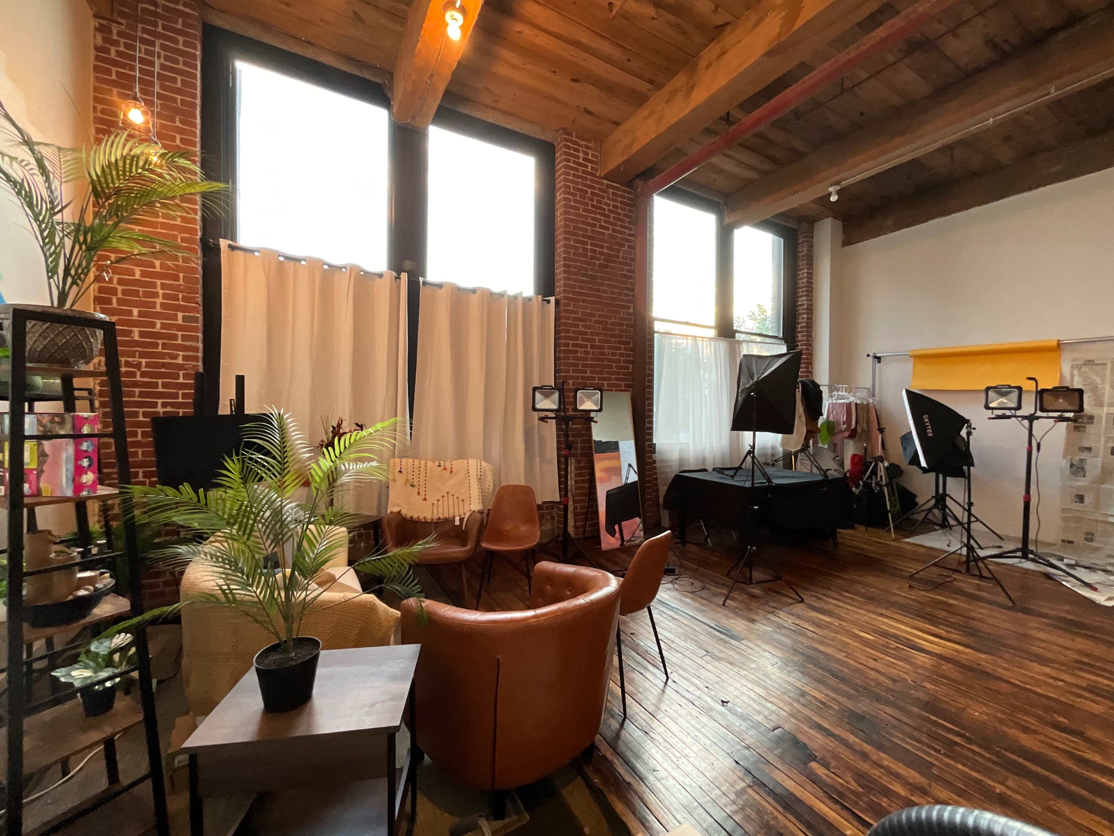 A spacious studio with brick walls, large windows, and various seating arrangements, surrounded by photography equipment and plants.