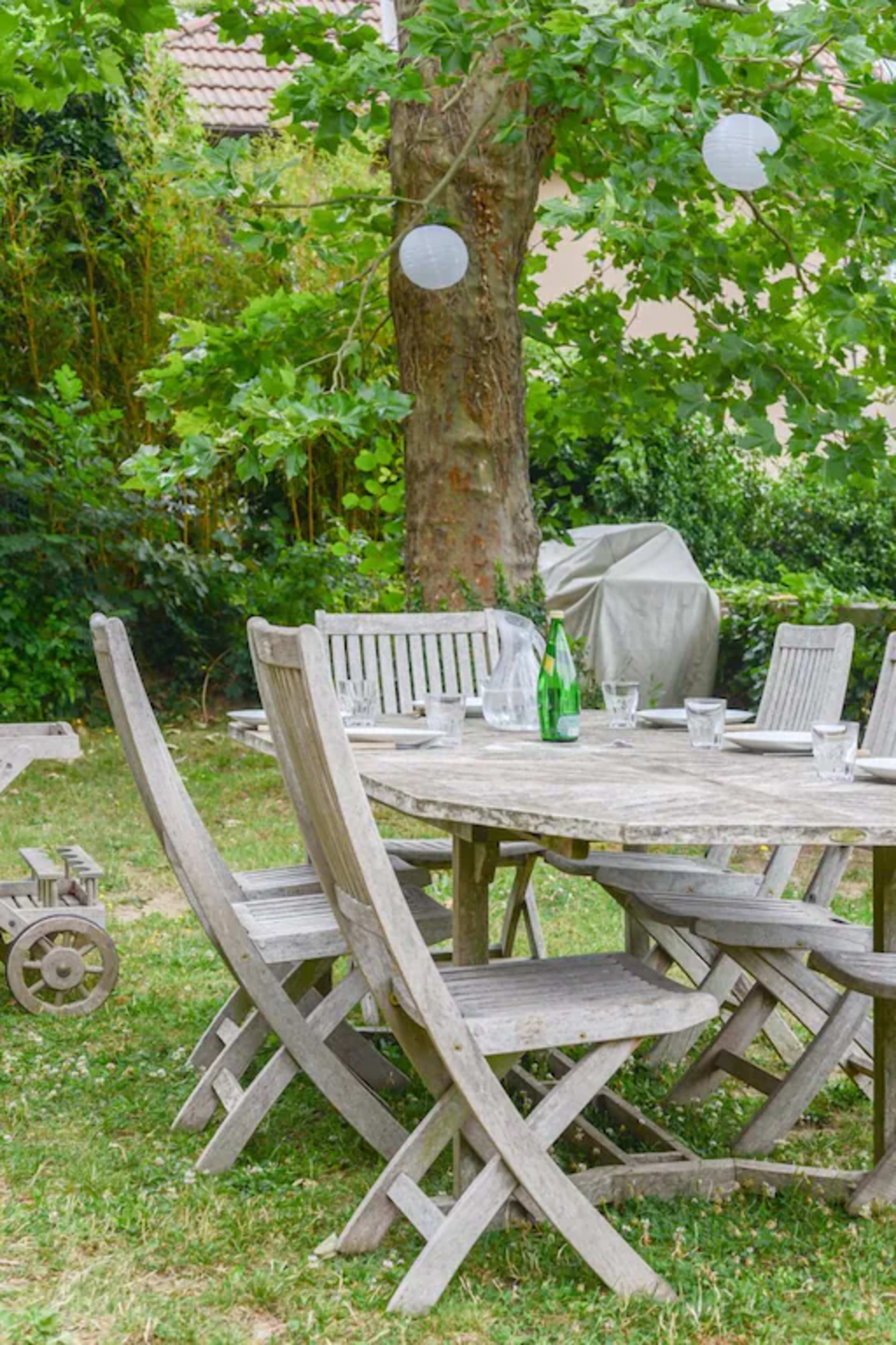 A weathered wooden dining table surrounded by folding chairs sits under a large tree in a green garden, with glassware and a bottle of water arranged on the table.