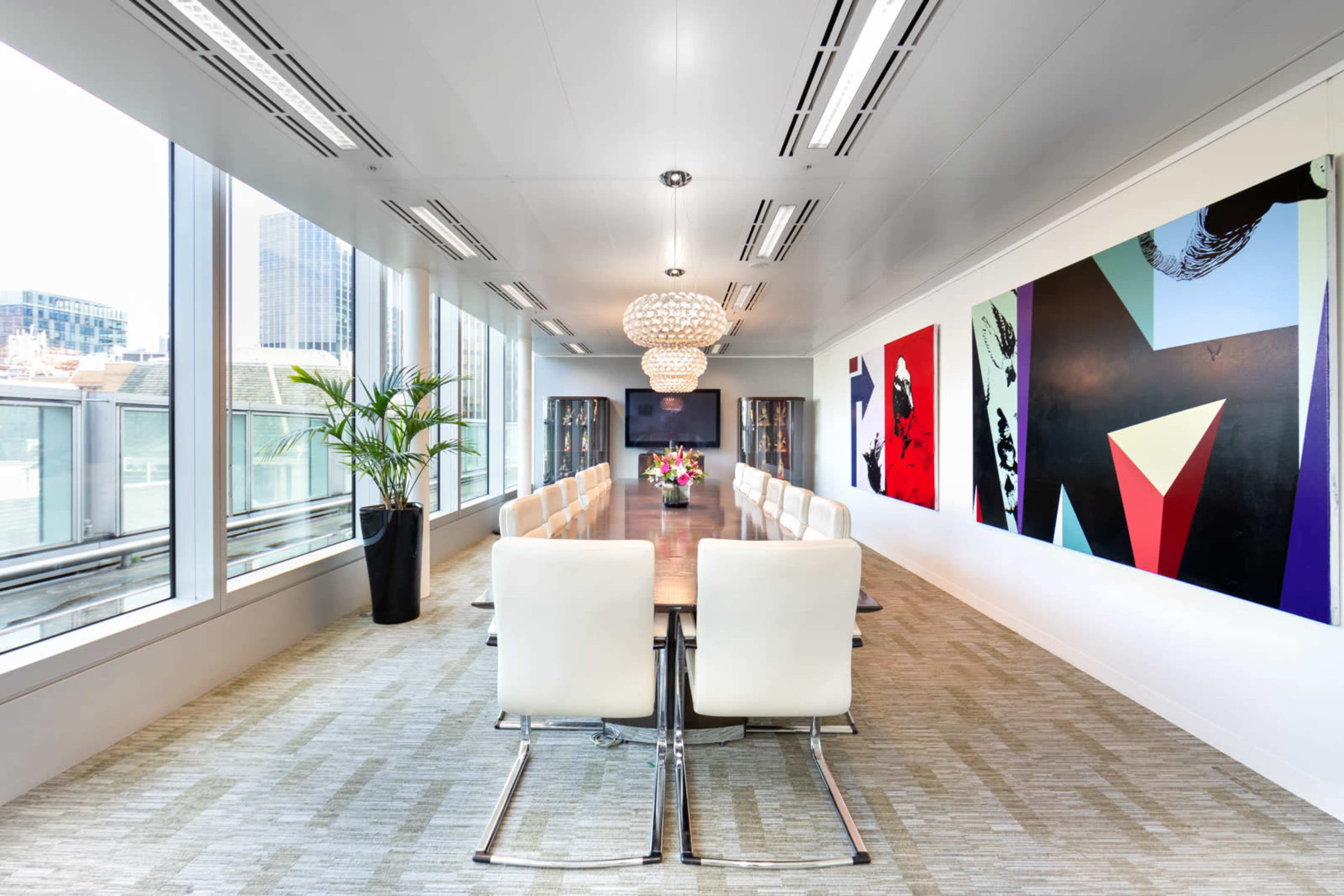 A long conference room features a large table surrounded by white chairs, with floor-to-ceiling windows on one side and colorful artwork on the walls.