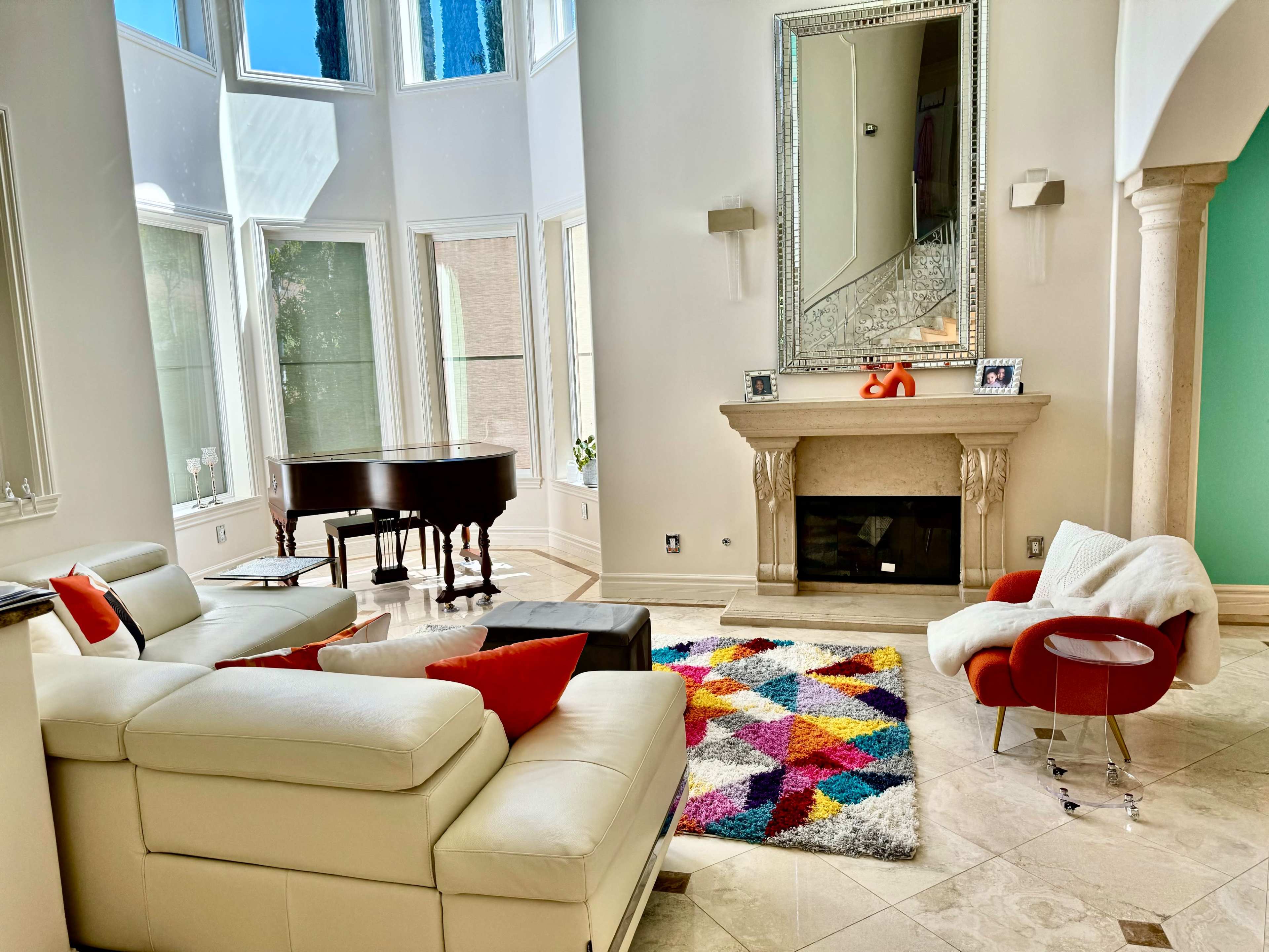 A spacious living room features a white leather sectional sofa, a colorful rug, a grand piano, and a fireplace beneath a large mirror.