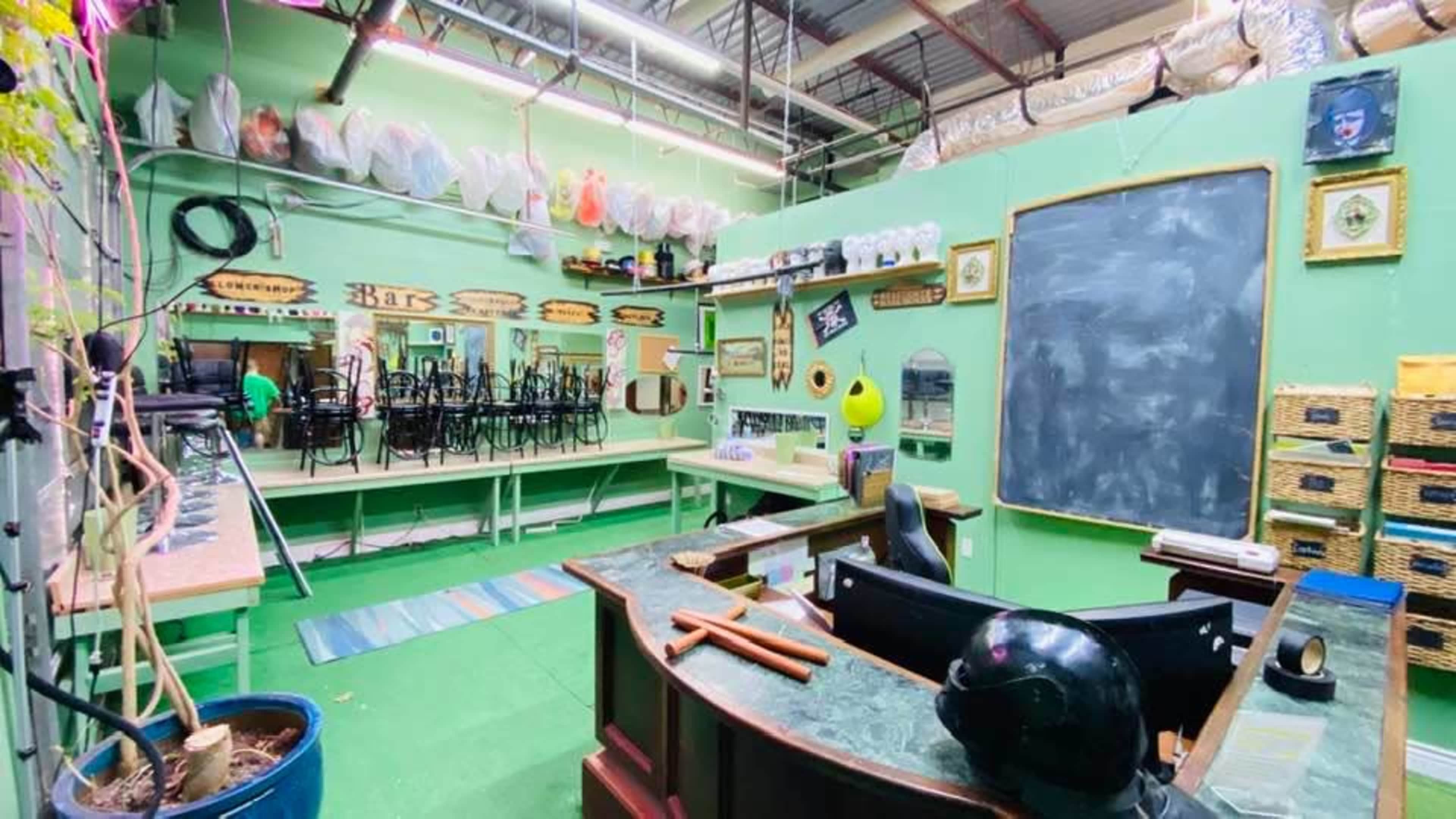 The image shows a brightly colored workshop space featuring a chalkboard, mirrors, shelves with supplies, and stacked chairs along the walls.