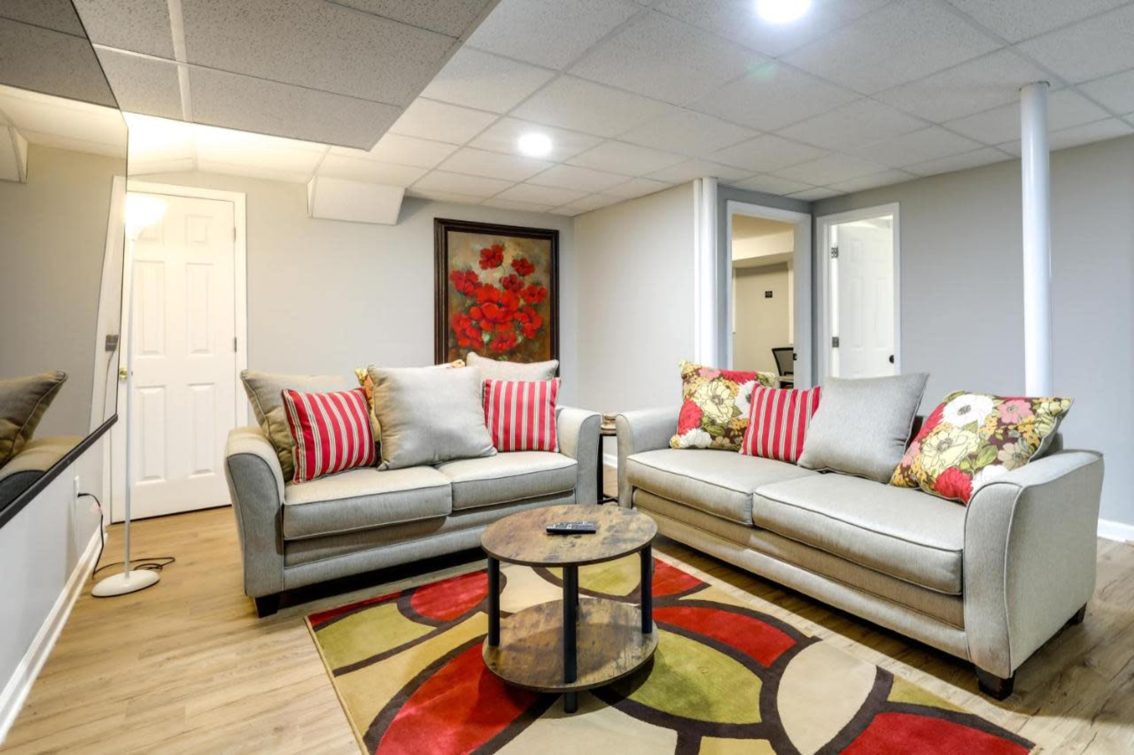 A basement living area features two sofas with decorative pillows, a round coffee table, and a colorful area rug under bright ceiling lights.