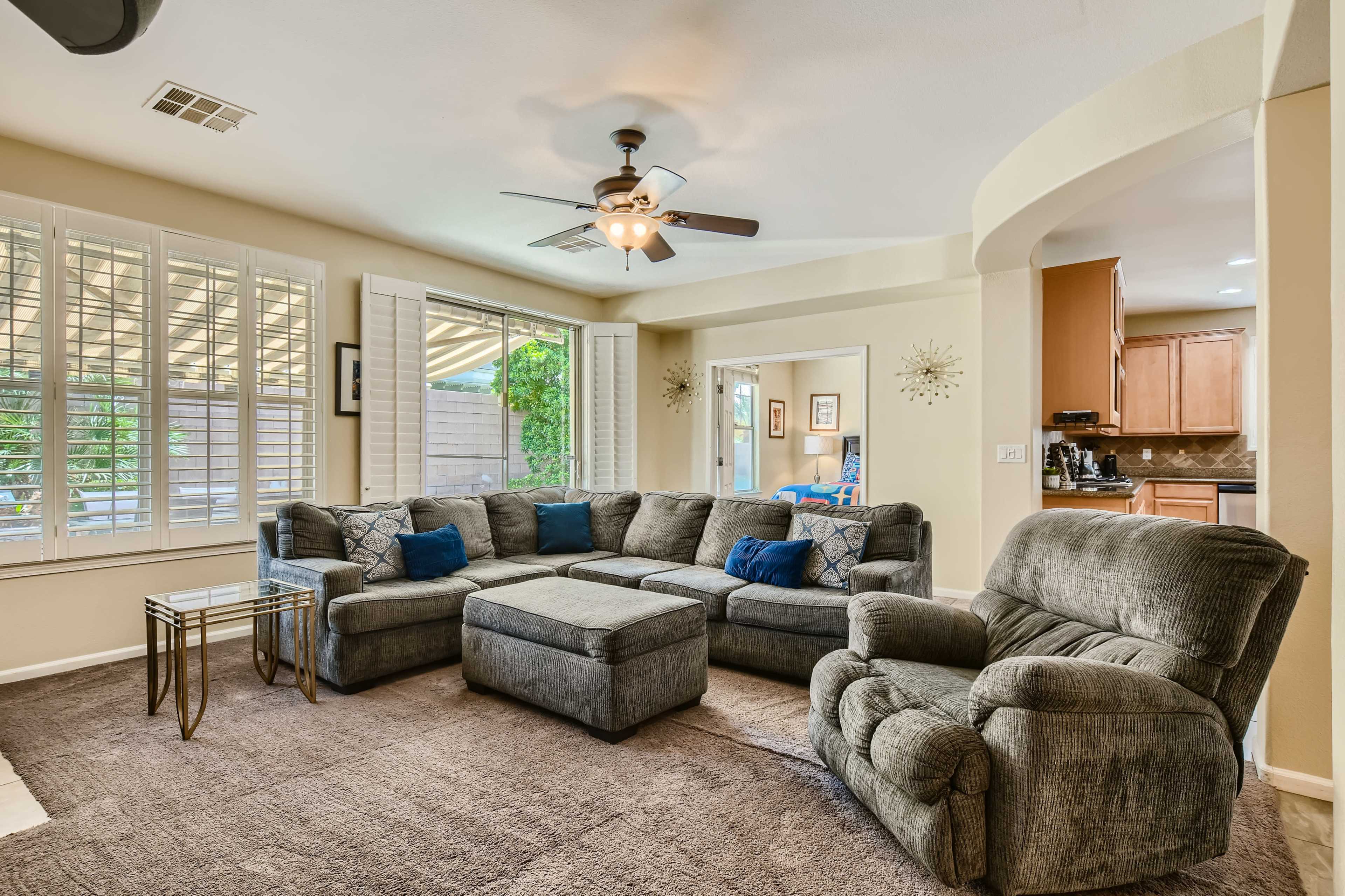A spacious living room features a large sectional sofa, a coffee table, and a ceiling fan, with light from windows illuminating the space.