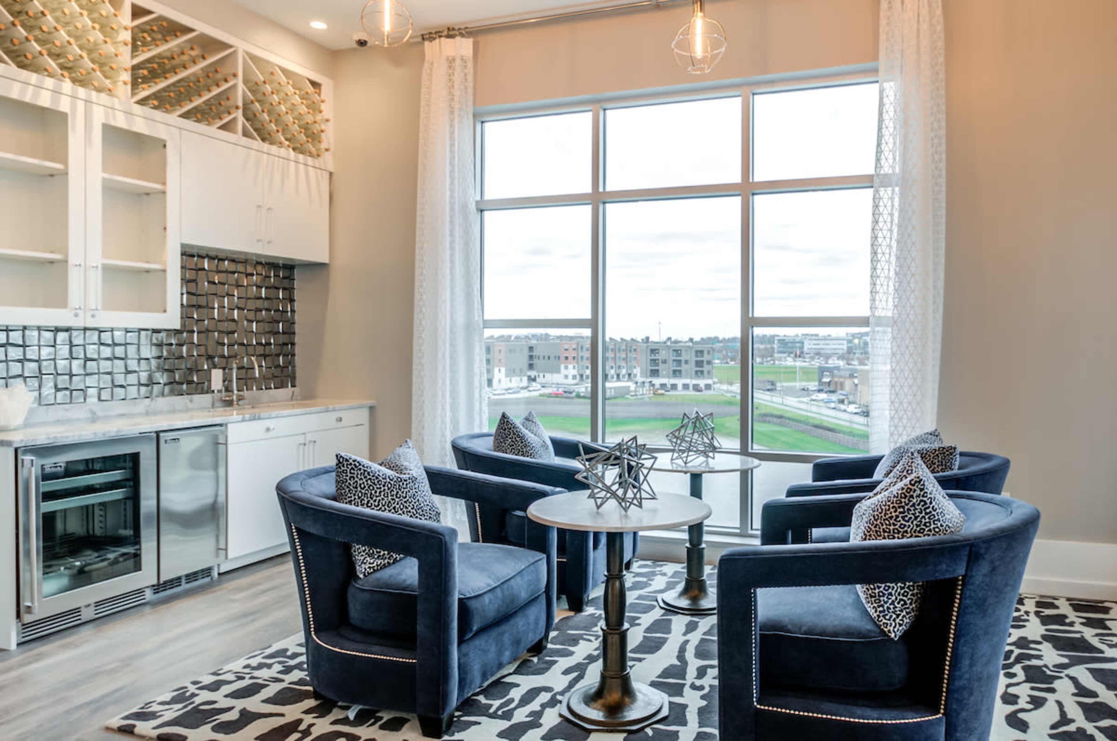 A modern living area with four blue armchairs arranged around a circular table, large windows offering a view of the outdoors, and a light-colored kitchen area with white cabinetry.