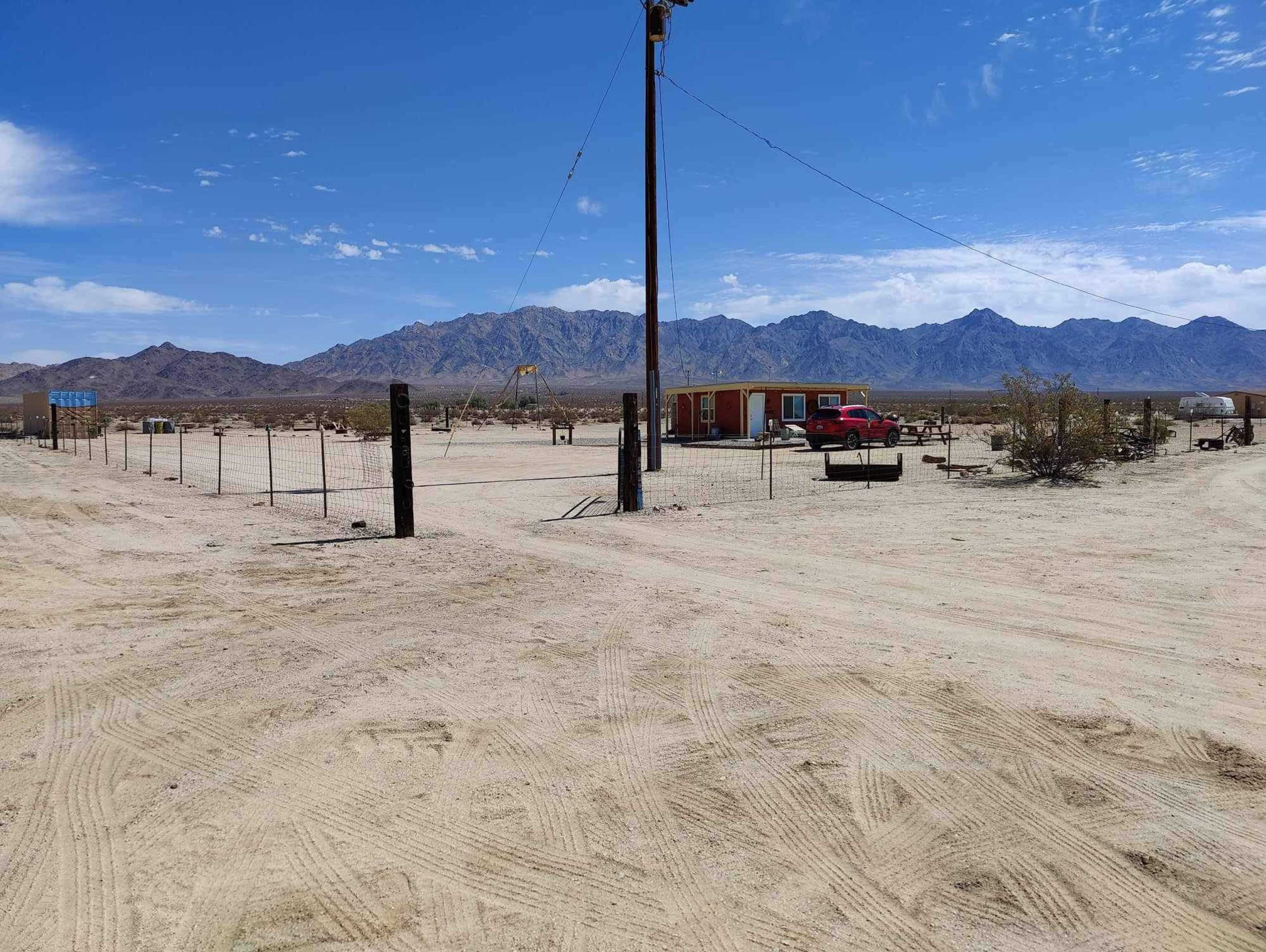 A barren desert landscape with a fenced area, a wooden structure, a red truck, and mountains in the background.