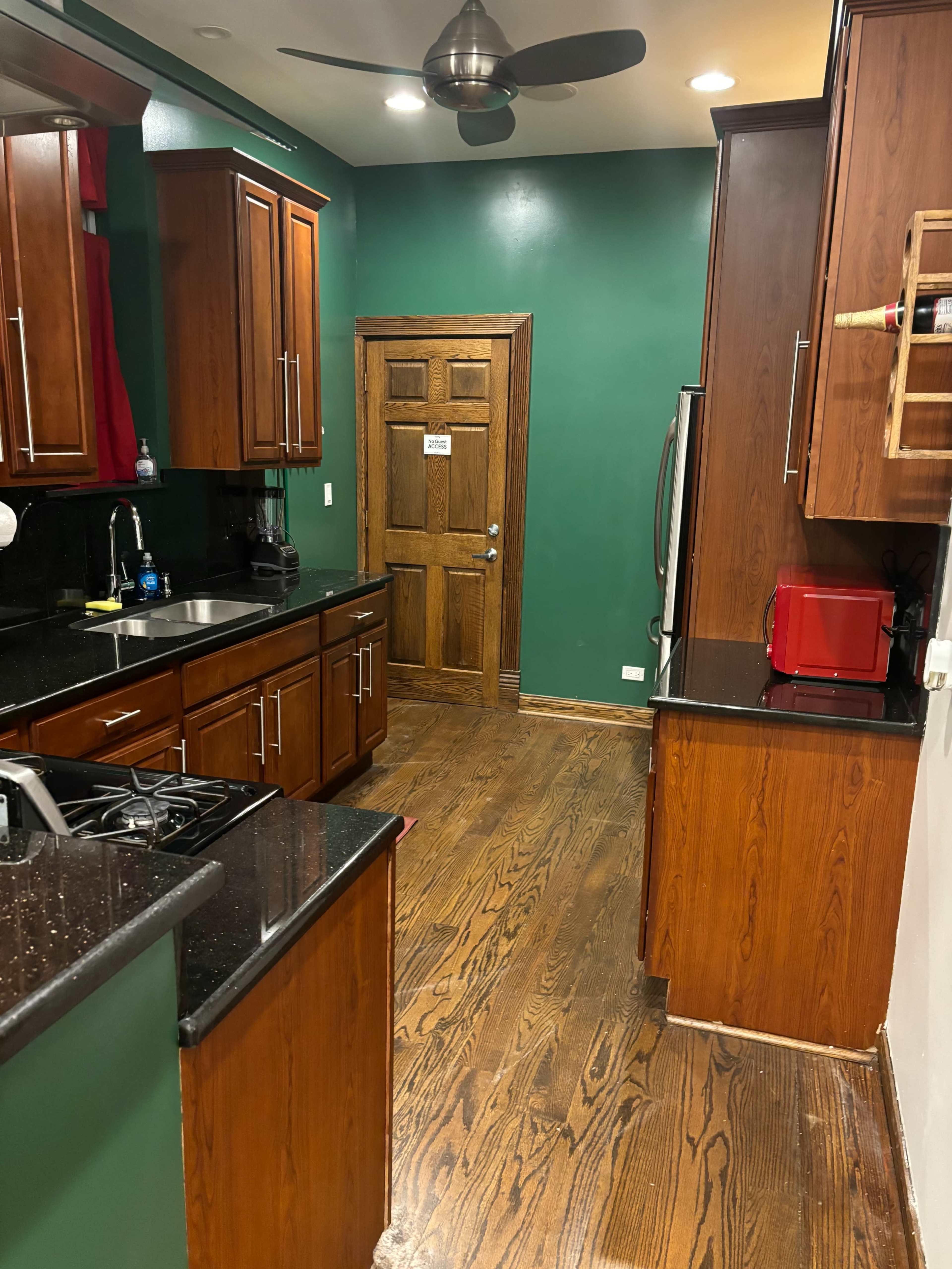 The image shows a kitchen with dark wooden cabinets and a green-painted wall, featuring a countertop and appliances on either side, leading to a door at the end of the room.