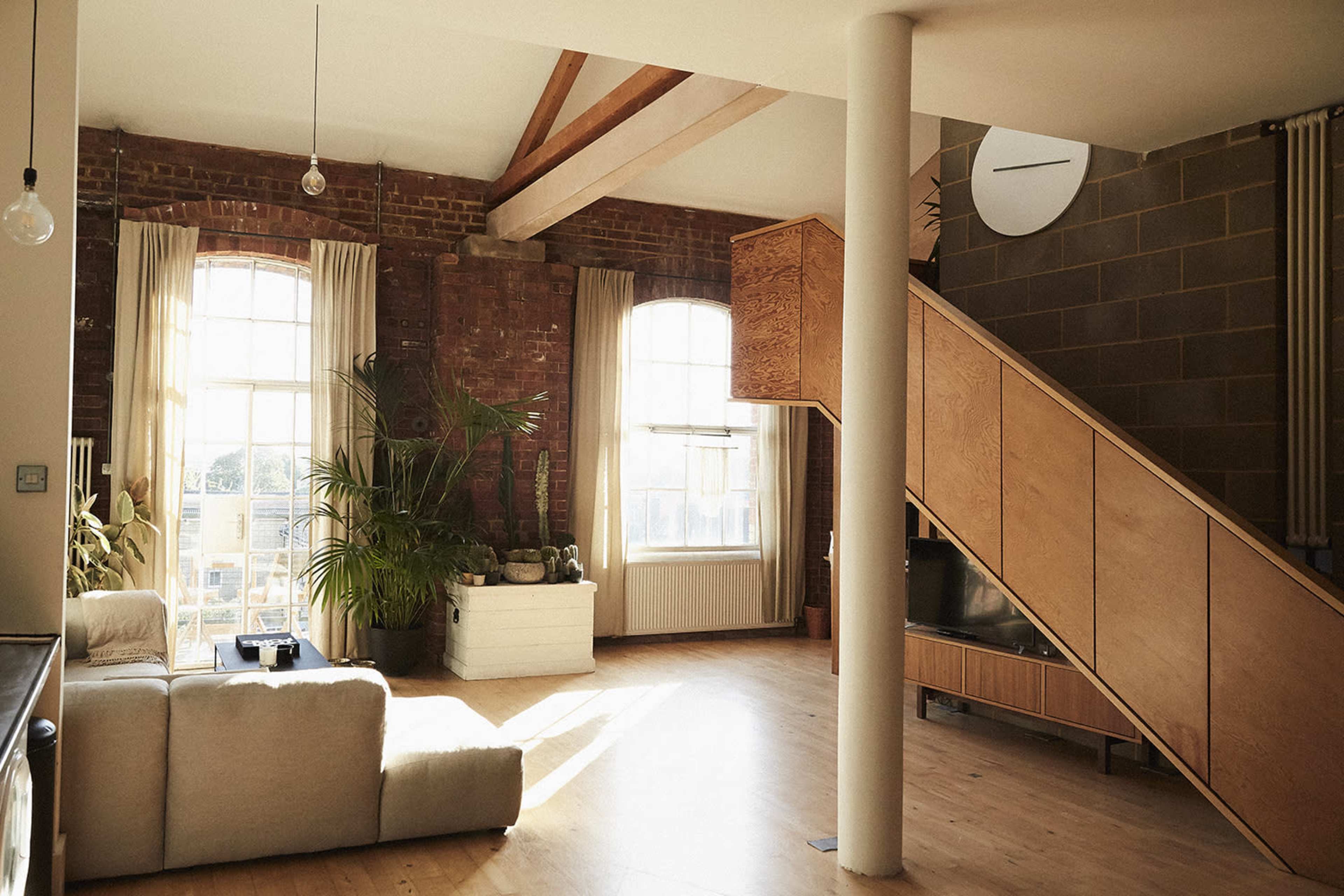 The image shows a modern living space featuring exposed brick walls, large windows, and a wooden staircase leading to an upper level.