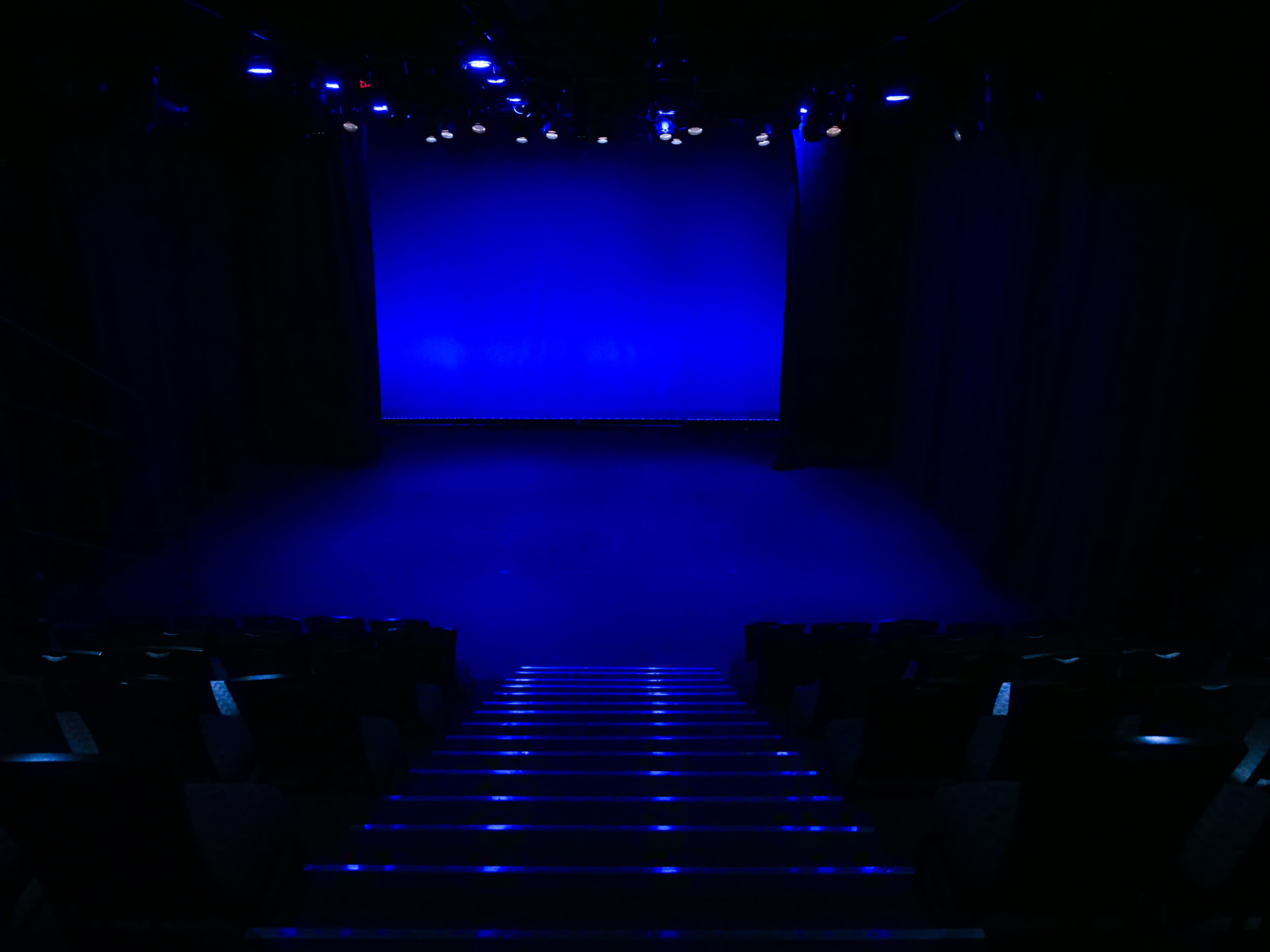 A dimly lit theater stage is visible, with blue lighting illuminating the backdrop and empty seating in the foreground.