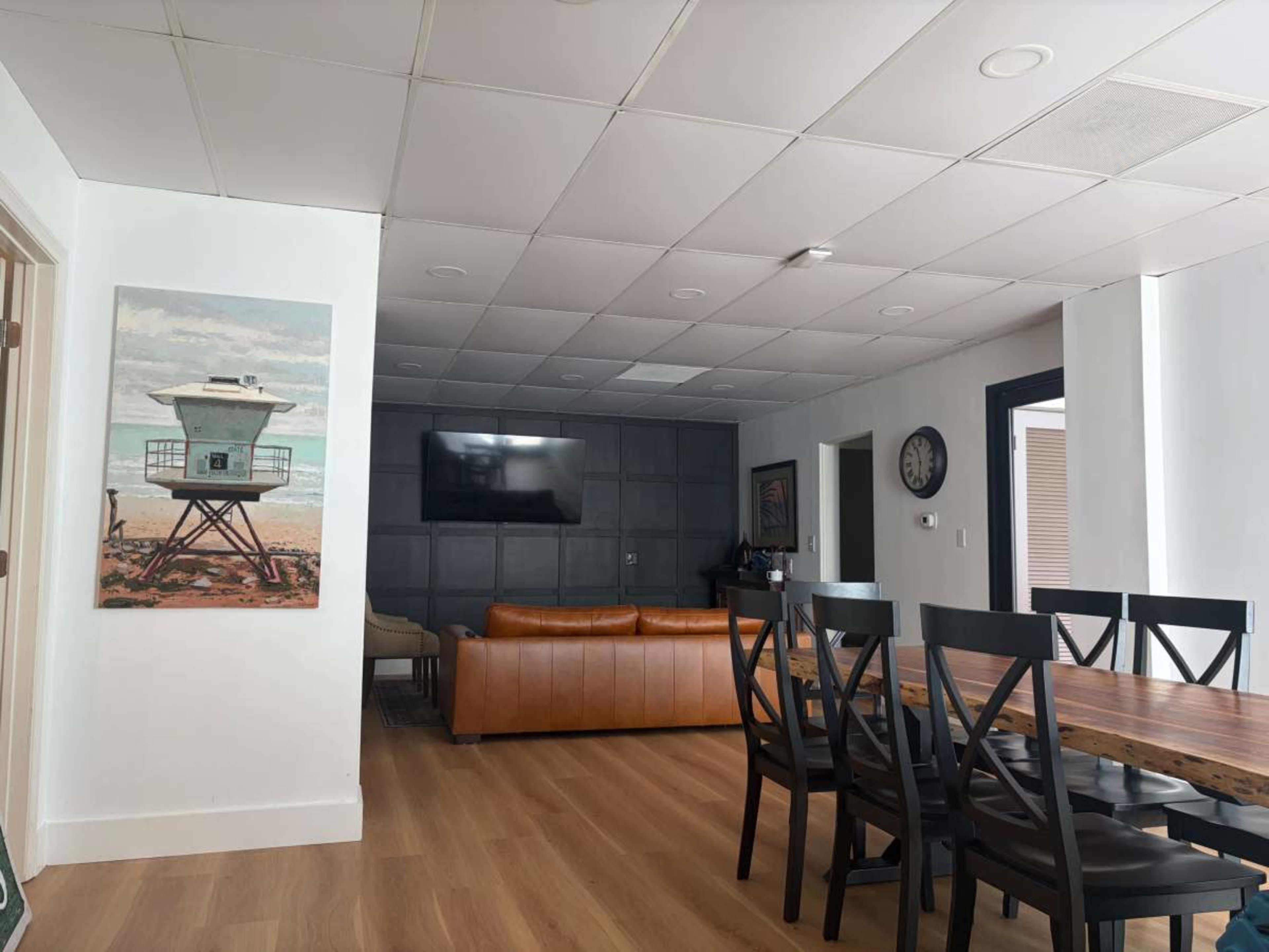 The image shows a modern, open living space featuring a leather sofa, a large wall-mounted TV, a wooden dining table, and a beach-themed painting.