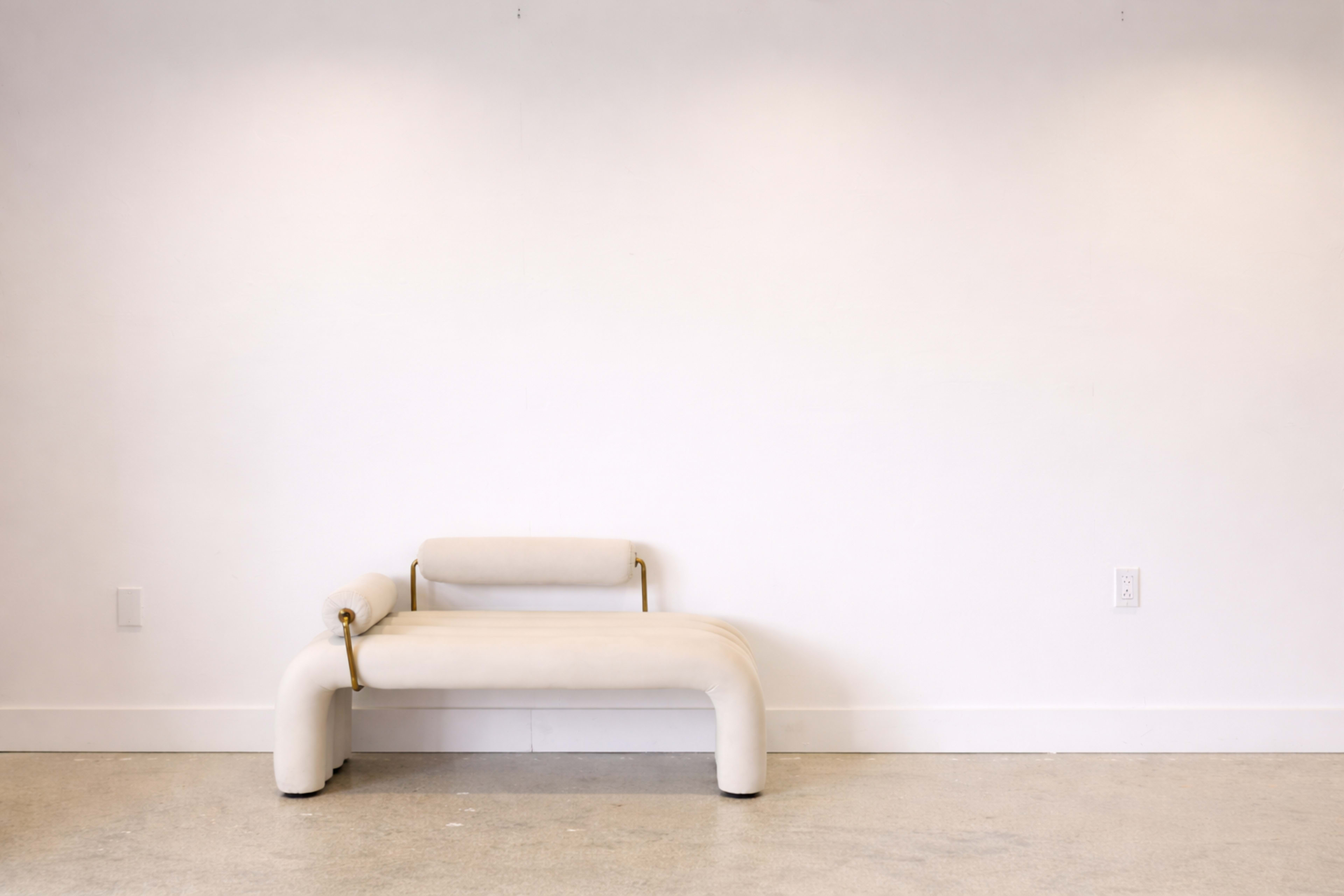 A curved, white bench with a cylindrical armrest is positioned against a plain white wall in a minimalistic room.